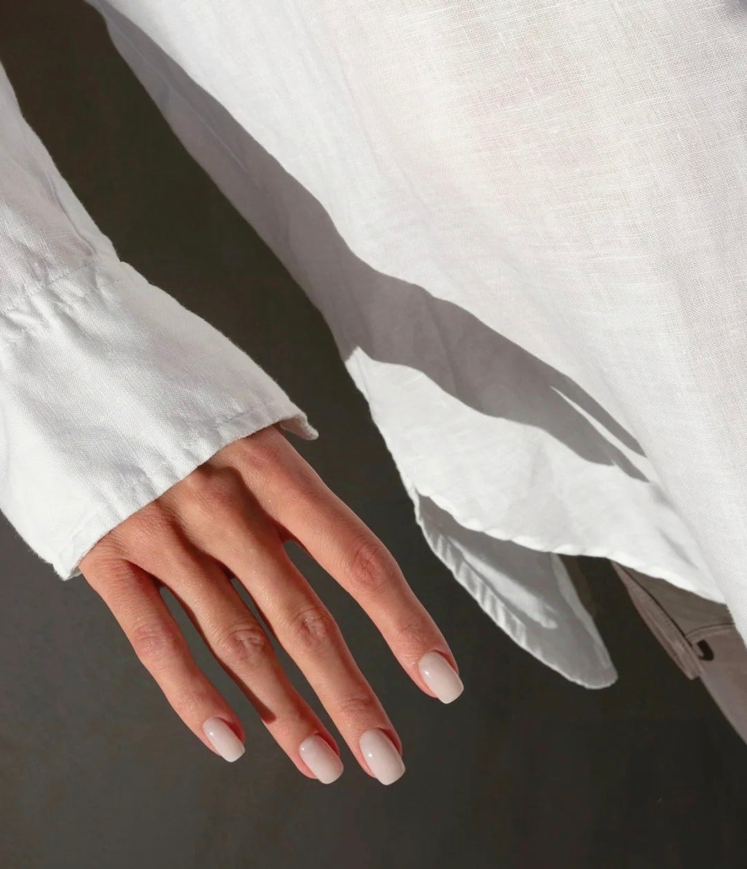 A close-up image of a woman's hand showcasing short oval nails in a natural nude shade with a subtle gloss. Her hand rests near the sleeve of a white linen shirt, highlighting the minimalist and elegant style of her manicure. Perfect for everyday wear, these nude nails offer a clean, sophisticated look.