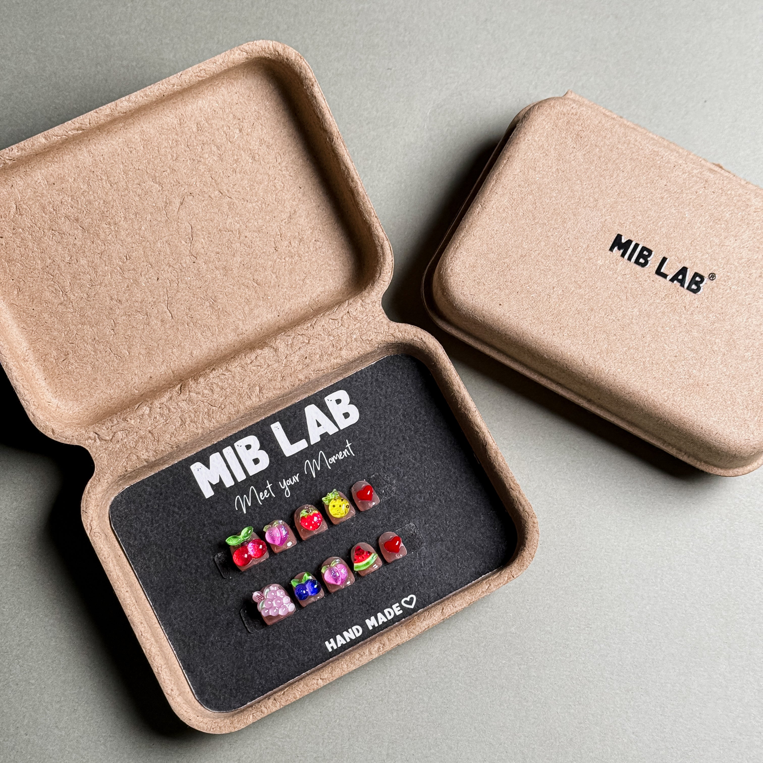 A set of Handmade press on nails with playful fruit and animal designs, presented in eco-friendly packaging. These Custom press on nails offer a fun and whimsical look, perfect for Everyday nails with a unique touch. The box, labeled "MIB LAB" and "Meet Your Moment", highlights the brand's dedication to creating personalized Nail art designs. Ideal for those seeking Reusable fake nails that combine style and creativity.