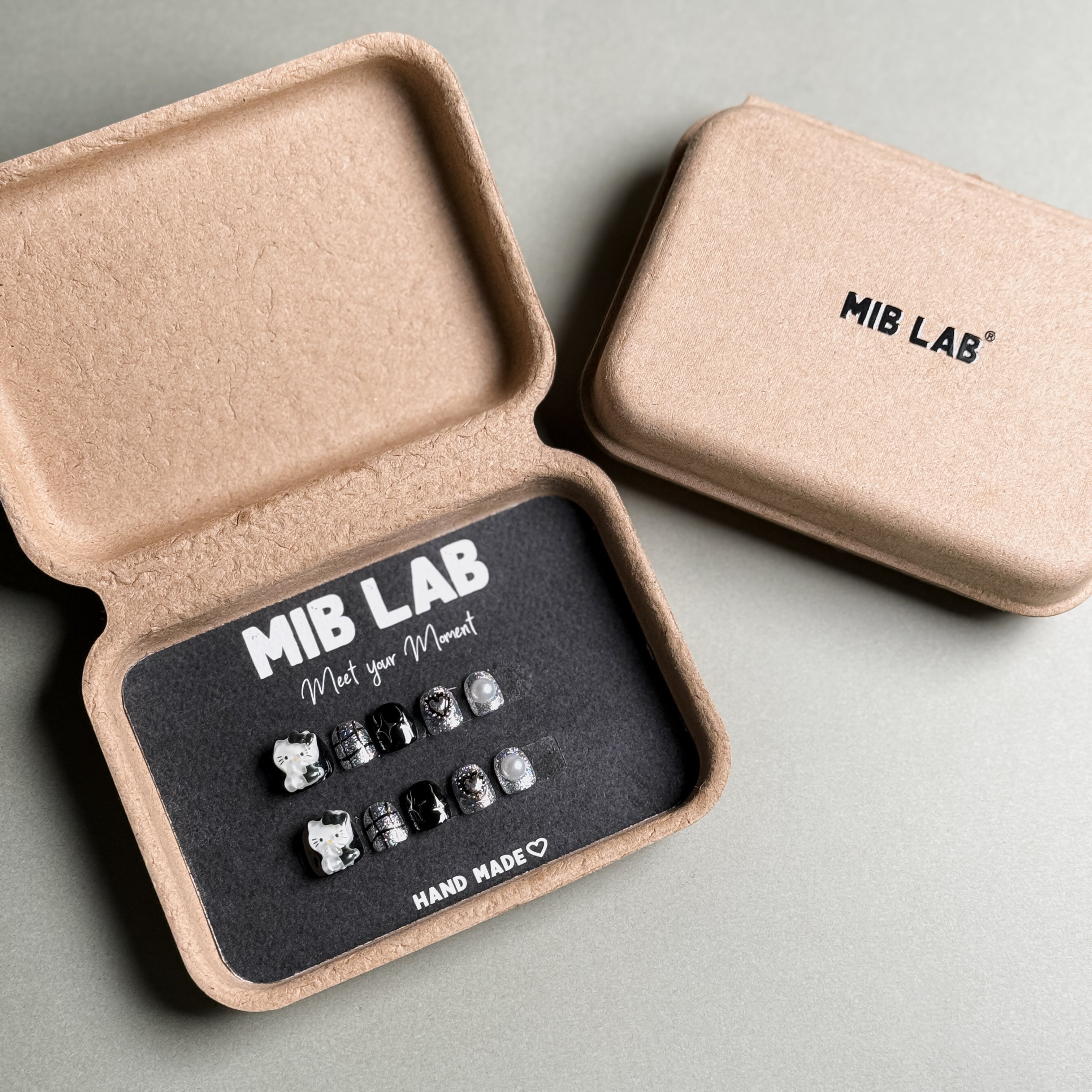 A set of Handmade press on nails with elegant pearl and silver accents, presented in eco-friendly packaging. These Luxury press on nails offer a sophisticated and timeless look, perfect for Wedding nails or special occasions. The box, labeled "MIB LAB" and "Meet Your Moment", highlights the brand's dedication to creating memorable Nail art designs. Ideal for those seeking Reusable fake nails that combine style and elegance.