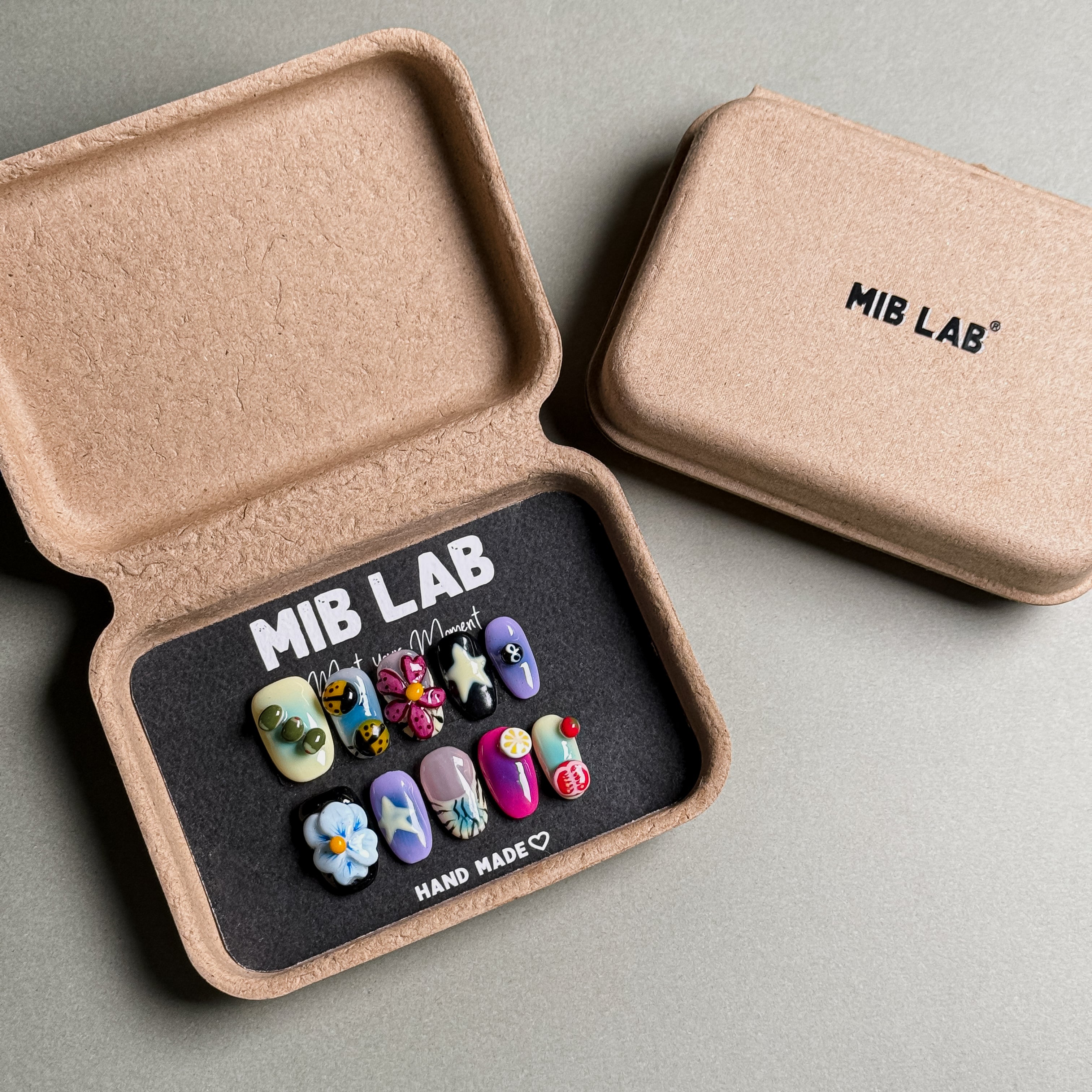 A set of Handmade press on nails with playful and whimsical designs, presented in eco-friendly packaging. These Custom press on nails offer a fun and unique touch, perfect for Everyday nails with a creative flair. The box, labeled "MIB LAB" and "Meet Your Moment", highlights the brand's dedication to creating personalized Nail art designs. Ideal for those seeking Reusable fake nails that combine style and sustainability.