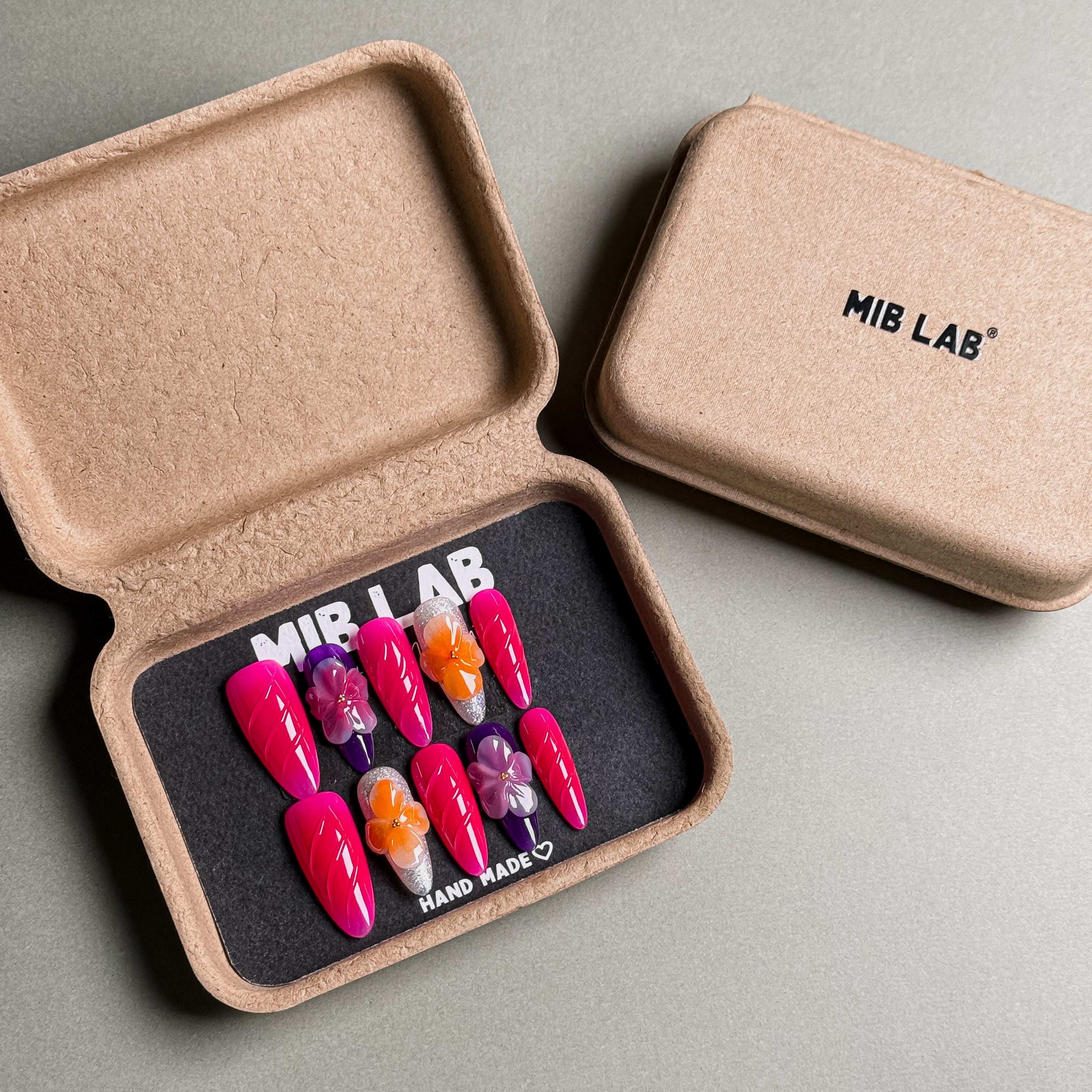 A set of Handmade press on nails with vibrant pink hues and floral accents, presented in eco-friendly packaging. These Custom press on nails offer a bold and feminine look, perfect for Party nails or special occasions. The box, labeled "MIB LAB" and "Meet Your Moment", highlights the brand's dedication to creating memorable Nail art designs. Ideal for those seeking Reusable fake nails that combine style and sustainability.