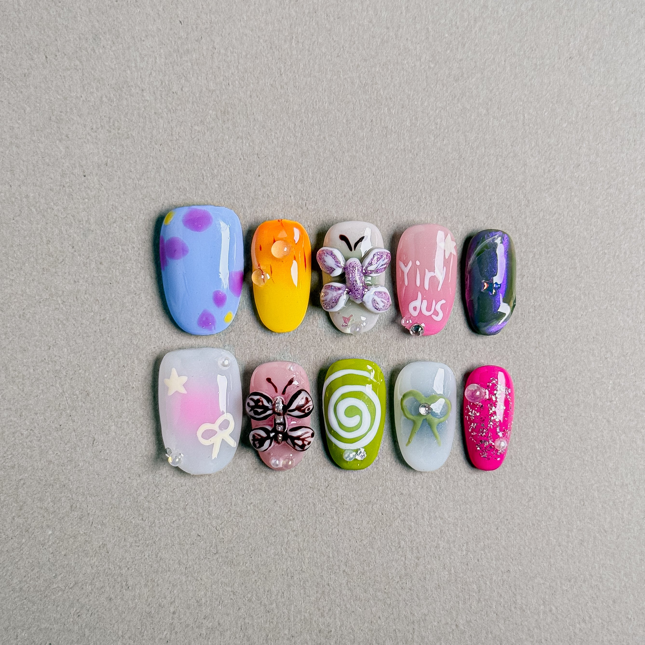 A set of "Bloom" press - on nails showcasing a variety of colorful and whimsical designs, including butterflies, swirls, and floral motifs, perfect for adding a fun and charming touch to daily or special - occasion nail looks.
