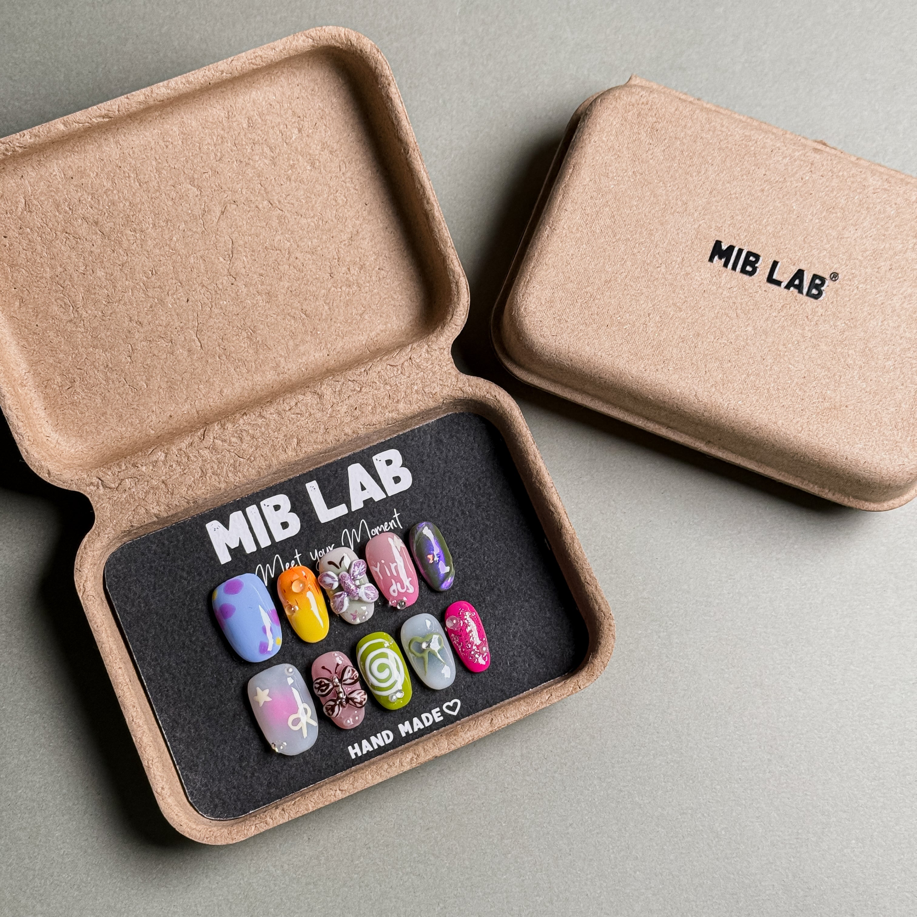 A set of Handmade press on nails with a variety of colorful and detailed designs, presented in eco-friendly packaging. These Custom press on nails offer a range of styles, perfect for Everyday nails with a unique touch. The box, labeled "MIB LAB" and "Meet Your Moment", highlights the brand's dedication to creating personalized Nail art designs. Ideal for those seeking Reusable fake nails that combine style and sustainability.