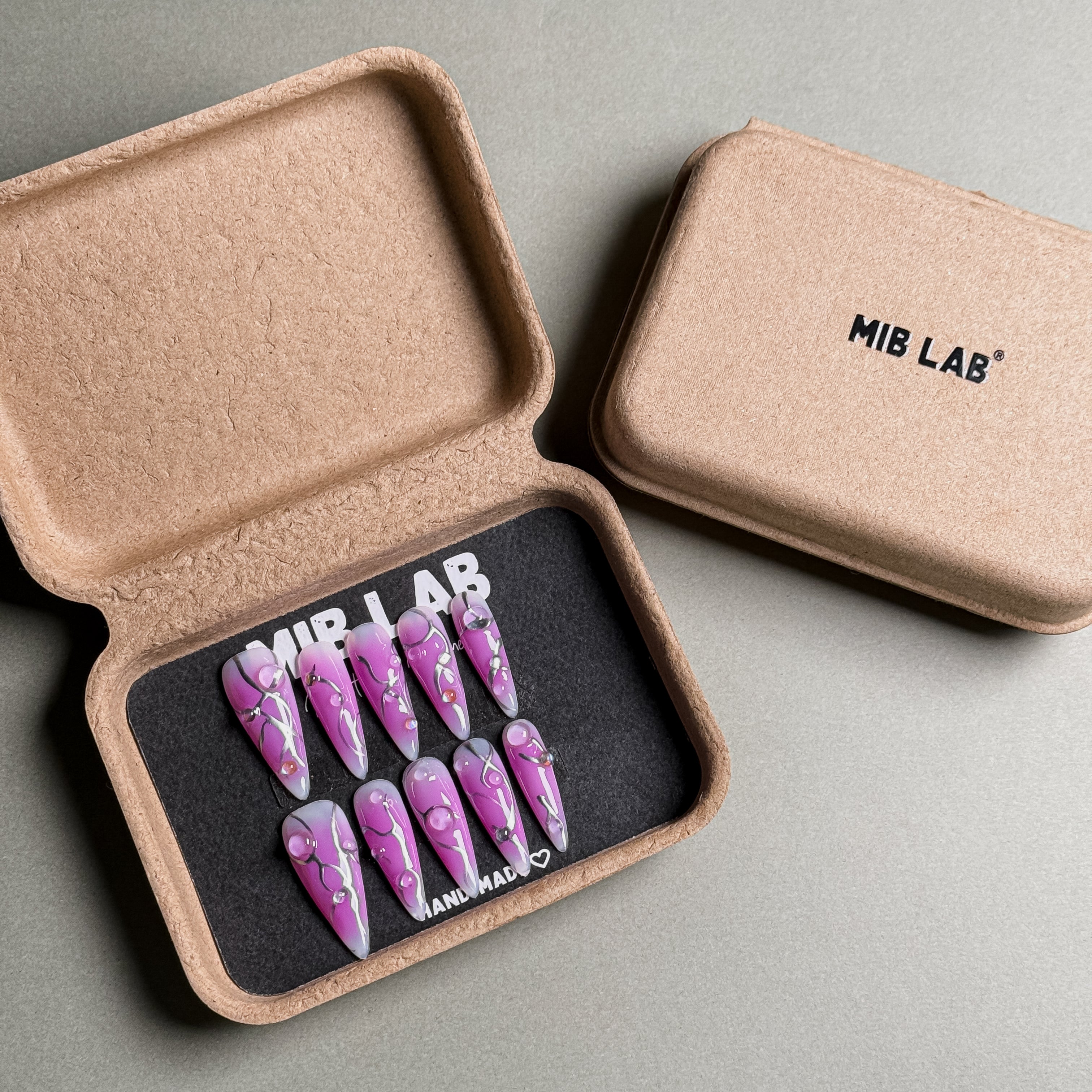 A set of Handmade press on nails with a vibrant purple design featuring abstract lines and silver accents, presented in eco-friendly packaging. These Custom press on nails offer a bold and artistic look, perfect for Party nails or special occasions. The box, labeled "MIB LAB" and "Meet Your Moment", highlights the brand's dedication to creating memorable Nail art designs. Ideal for those seeking Reusable fake nails that combine style and sustainability.