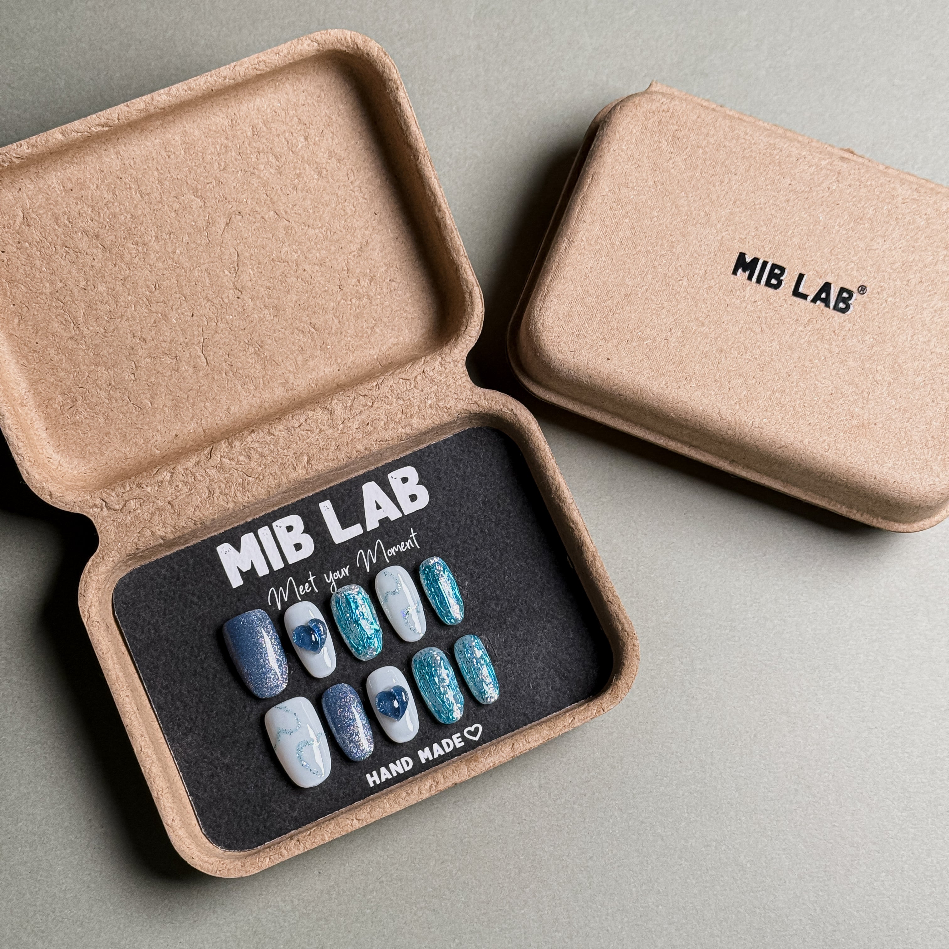 A set of Handmade press on nails with a shimmery blue and white design, presented in eco-friendly packaging. These Glitter nails offer a sparkling and eye-catching look, perfect for Party nails or special occasions. The box, labeled "MIB LAB" and "Meet Your Moment", highlights the brand's dedication to creating memorable Nail art designs. Ideal for those seeking Reusable fake nails that combine style and sustainability.