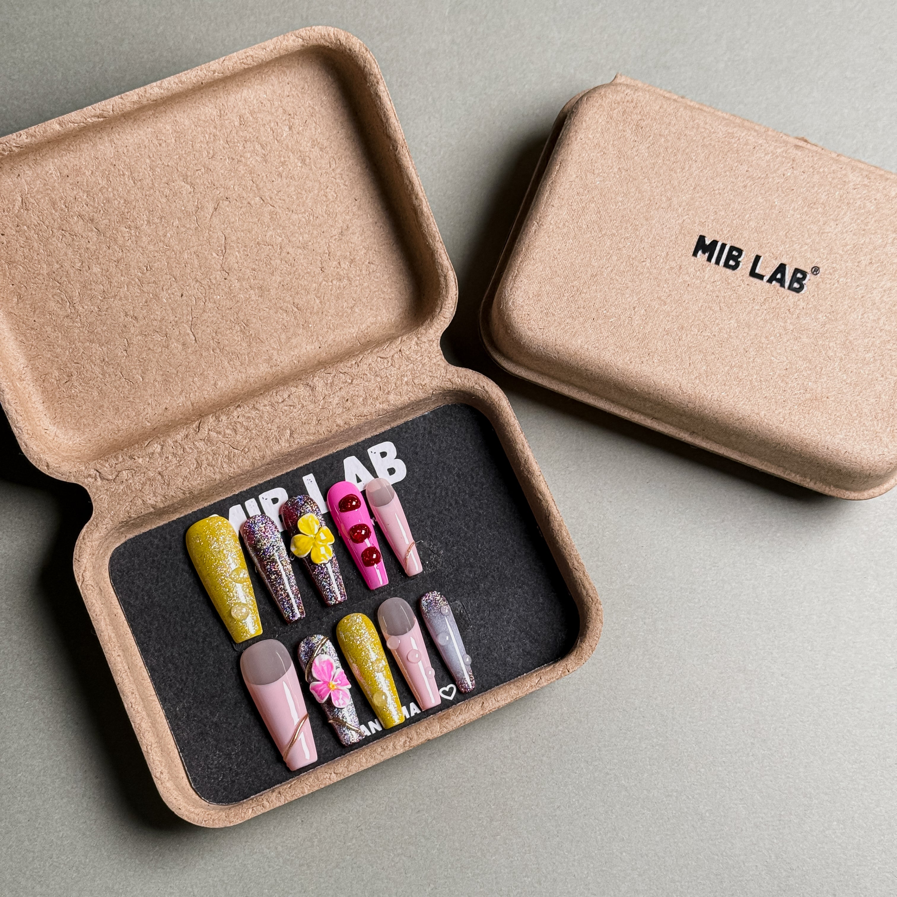 A set of Handmade press on nails with diverse designs, including floral accents and glitter details, presented in eco-friendly packaging. These Custom press on nails offer a variety of styles, perfect for Everyday nails with a unique touch. The box, labeled "MIB LAB" and "Meet Your Moment", highlights the brand's dedication to creating personalized Nail art designs. Ideal for those seeking Reusable fake nails that combine style and sustainability.