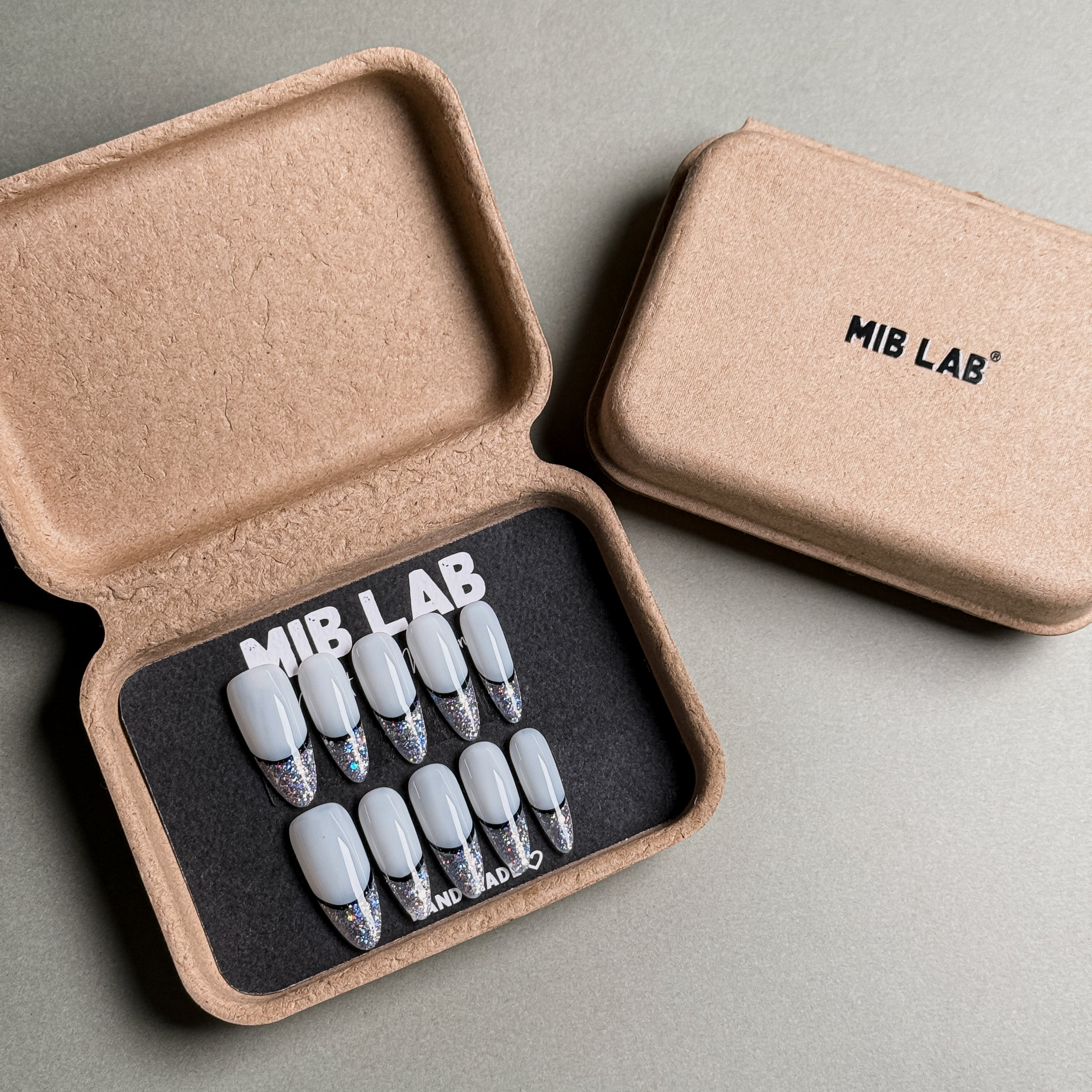 A set of Handmade press on nails with a classic French tip design and silver glitter accents, presented in eco-friendly packaging. These French tip nails offer a timeless and elegant look, perfect for Wedding nails or everyday wear. The box, labeled "MIB LAB" and "Meet Your Moment", highlights the brand's dedication to creating memorable Nail art designs. Ideal for those seeking Reusable fake nails that combine style and sophistication.