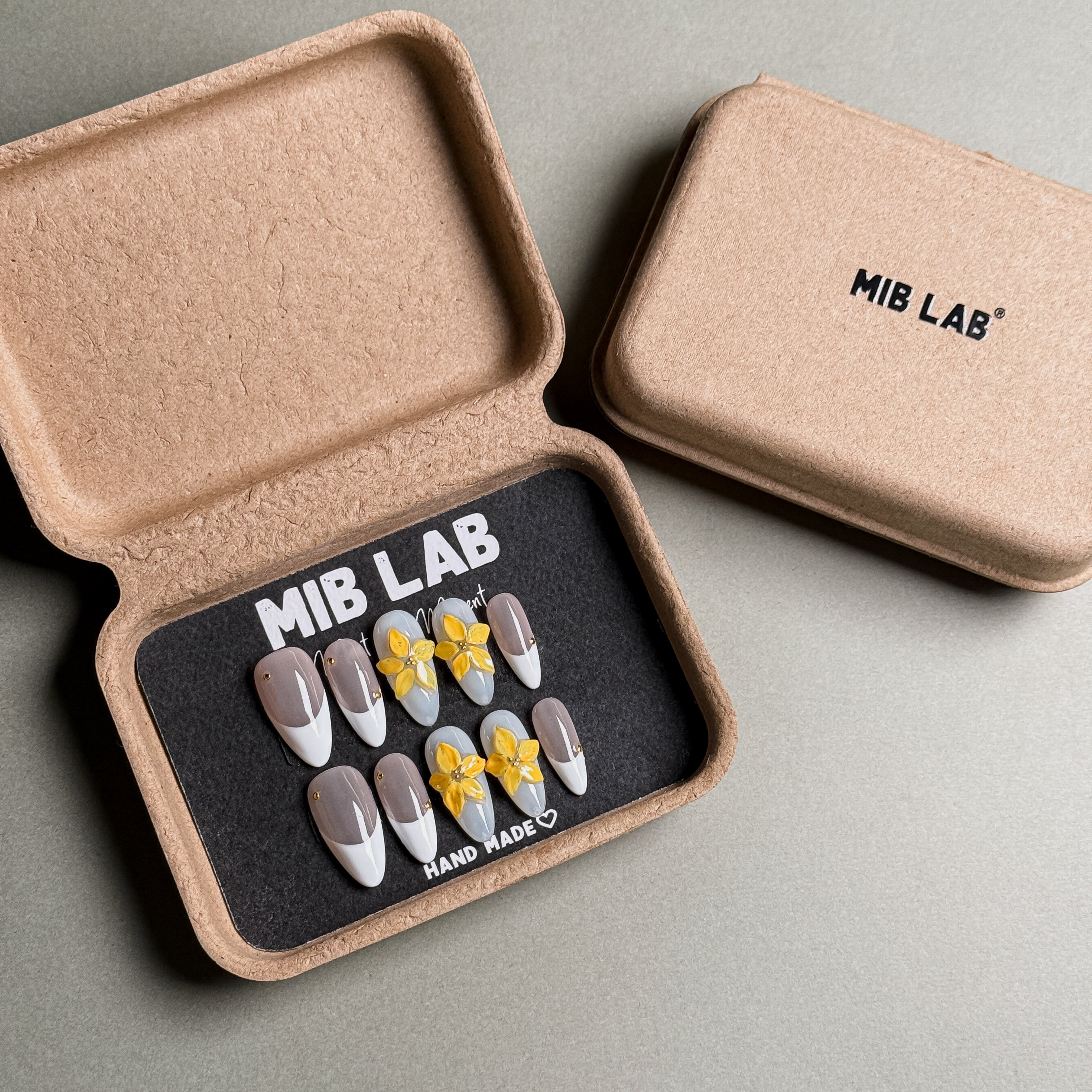 A set of Handmade press on nails with a classic French tip design and delicate yellow flower accents, presented in eco-friendly packaging. These French tip nails offer a timeless and elegant look, perfect for Wedding nails or everyday wear. The box, labeled "MIB LAB" and "Meet Your Moment", highlights the brand's dedication to creating memorable Nail art designs. Ideal for those seeking Reusable fake nails that combine style and sophistication.