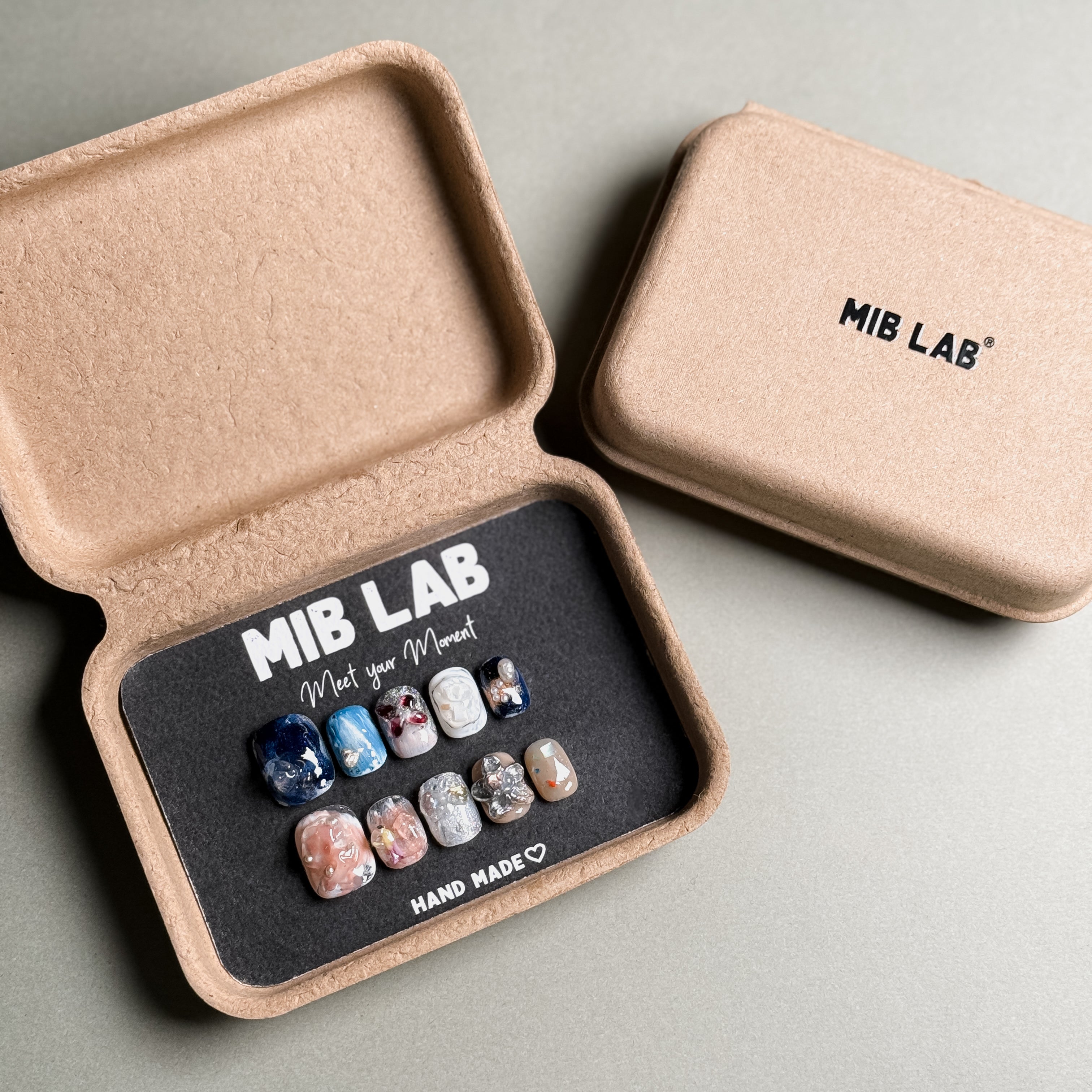 A collection of Handmade press on nails with diverse designs, including marble, glitter, and abstract patterns, presented in eco-friendly packaging. These Custom press on nails offer a variety of styles, perfect for Everyday nails with a unique touch. The box, labeled "MIB LAB" and "Meet Your Moment", highlights the brand's dedication to creating personalized Nail art designs. Ideal for those seeking Reusable fake nails that combine style and sustainability.