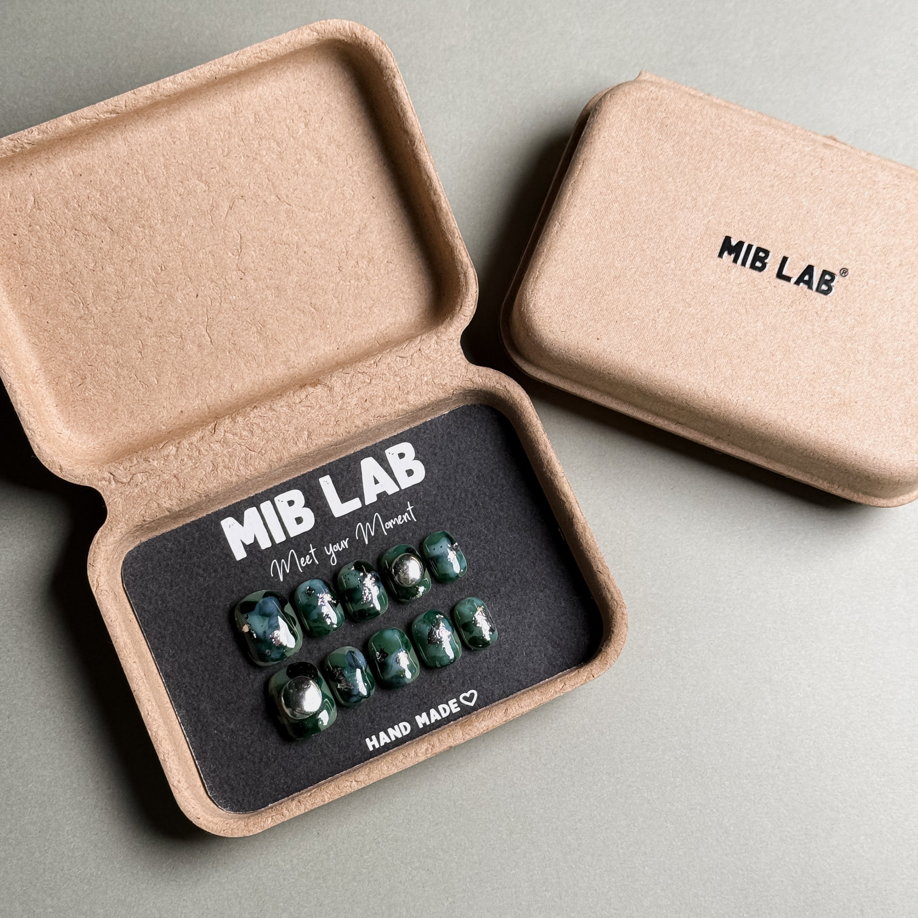A set of Handmade press on nails with a deep green, iridescent design, presented in eco-friendly packaging. These Luxury press on nails offer a bold and unique look, perfect for Party nails or special occasions. The box, labeled "MIB LAB" and "Meet Your Moment", highlights the brand's dedication to creating memorable Nail art designs. Ideal for those seeking Reusable fake nails that make a statement.