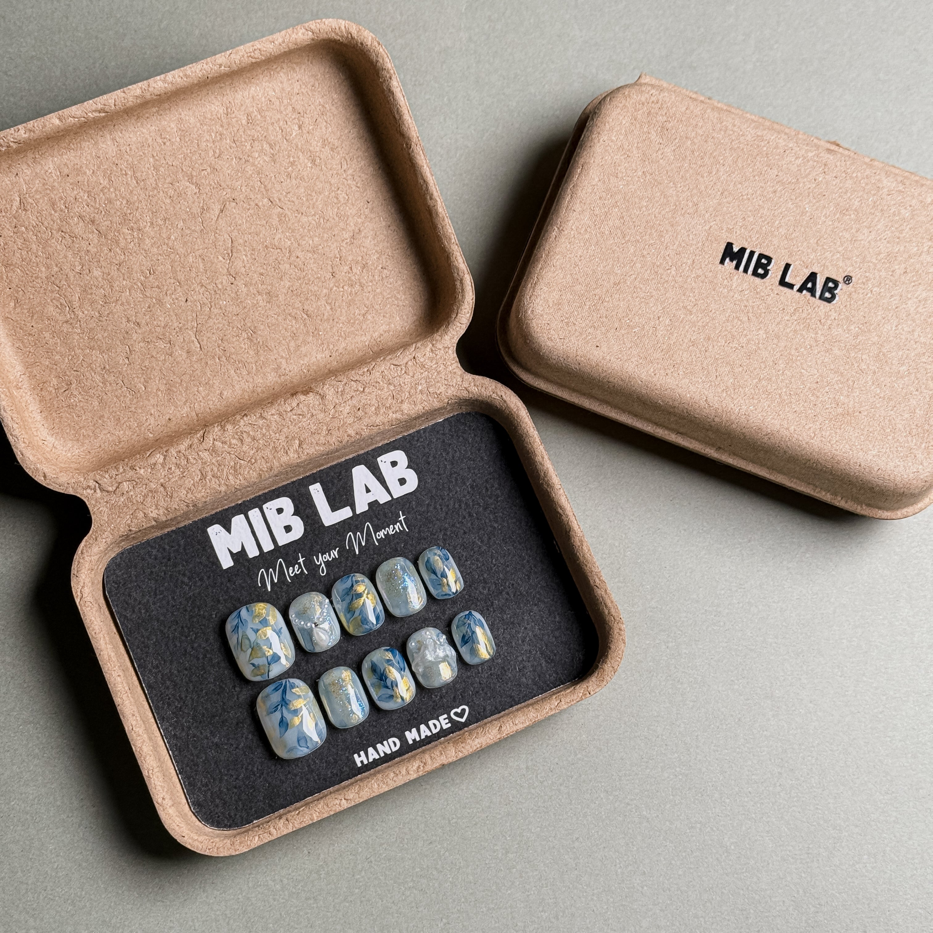 A set of Handmade press on nails with a delicate floral design on a light blue base, presented in eco-friendly packaging. These Floral nails offer a fresh and elegant look, perfect for Everyday nails with a touch of sophistication. The box, labeled "MIB LAB" and "Meet Your Moment", highlights the brand's dedication to creating memorable Nail art designs. Ideal for those seeking Reusable fake nails that combine style and sustainability.