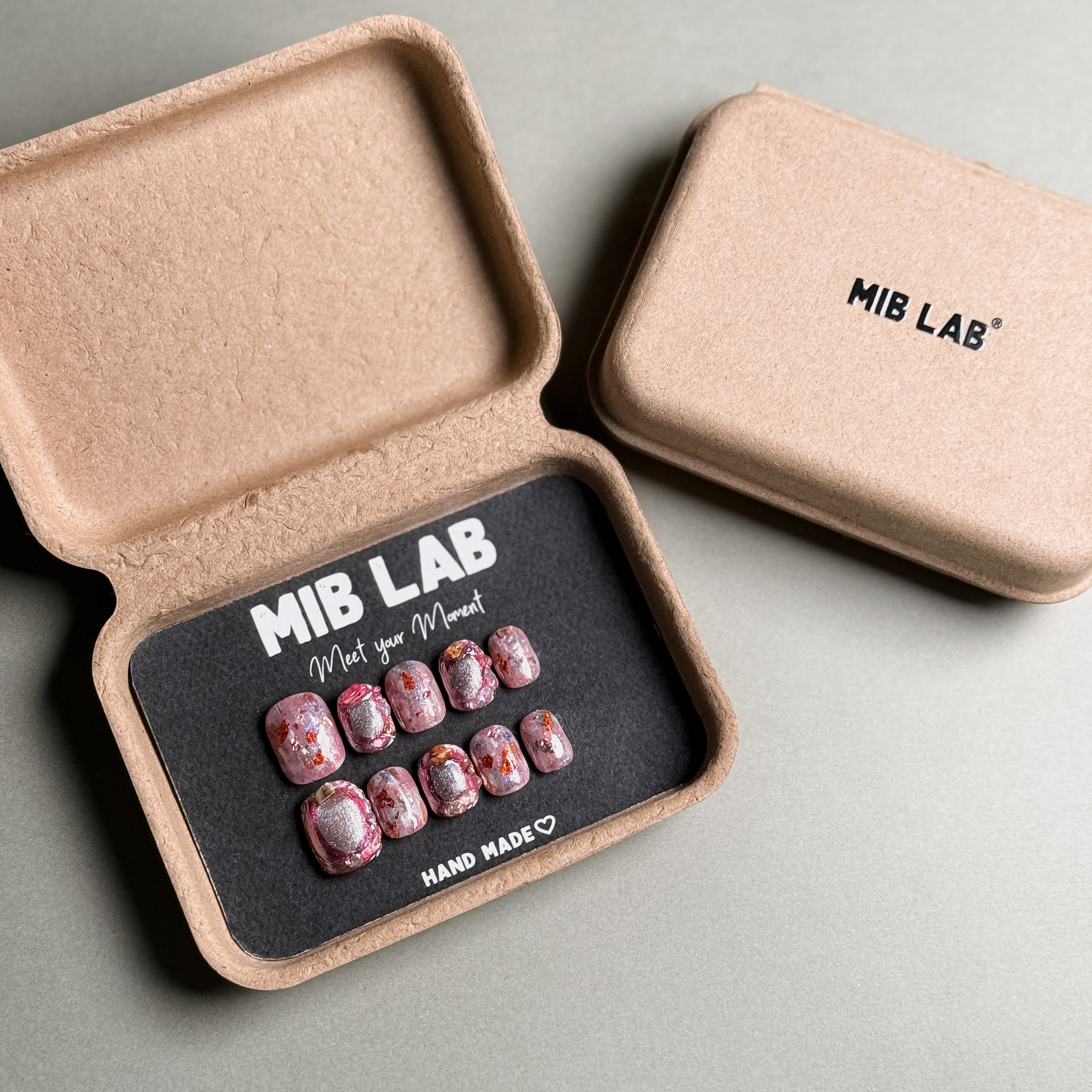A collection of Handmade press on nails with pink and glitter accents, neatly arranged in eco-friendly packaging. These Luxury press on nails feature intricate designs, perfect for a special occasion or as Wedding nails. The box, labeled "MIB LAB" and "Meet Your Moment", highlights the brand's dedication to creating memorable Nail art designs. Ideal for those seeking Reusable fake nails that combine elegance with sustainability.