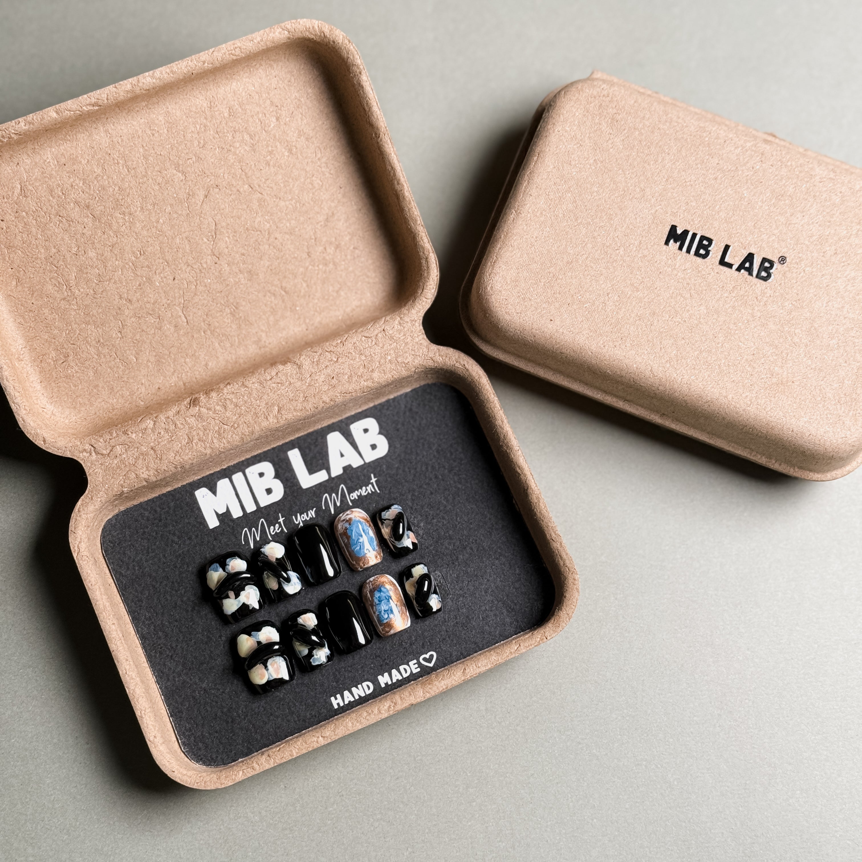 MIBLAB brand press-on fake nail set, featuring black short nails with abstract color patterns and 3D black decorations, handcrafted, packaged in an eco-friendly recycled paper box, showcasing the brand name "MIB LAB" and slogan "Meet your Moment."