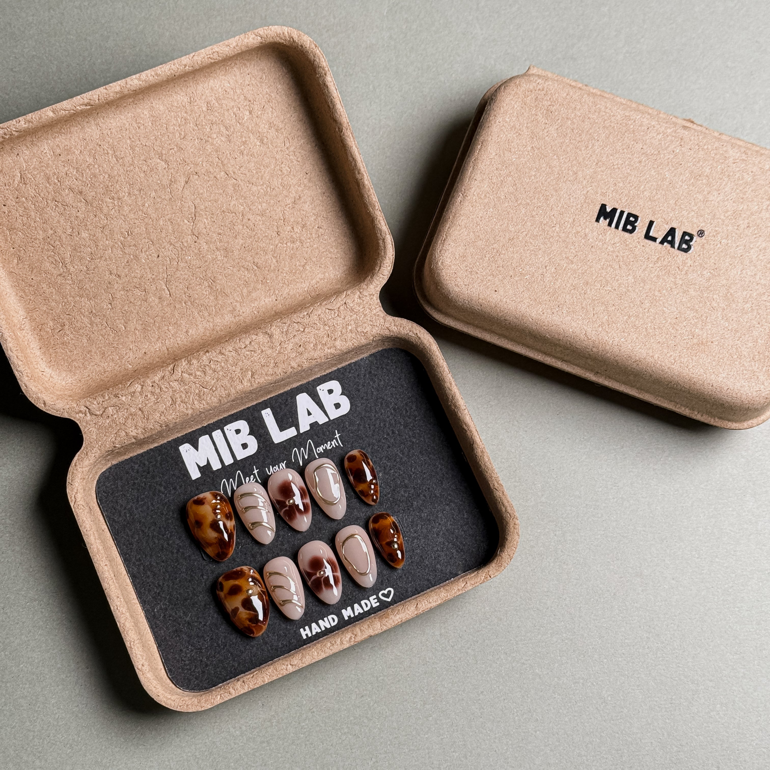 A set of handmade press-on nails with unique tortoise shell and gold foil designs, housed in eco-friendly packaging. These custom press-on nails offer a sophisticated and artistic look, perfect for an everyday manicure with a touch of luxury. The box is labeled "MIB LAB" and "Meet Your Moment," underscoring the brand's commitment to creating personalized nail designs. Perfect for those seeking a luxurious press-on manicure that stands out.