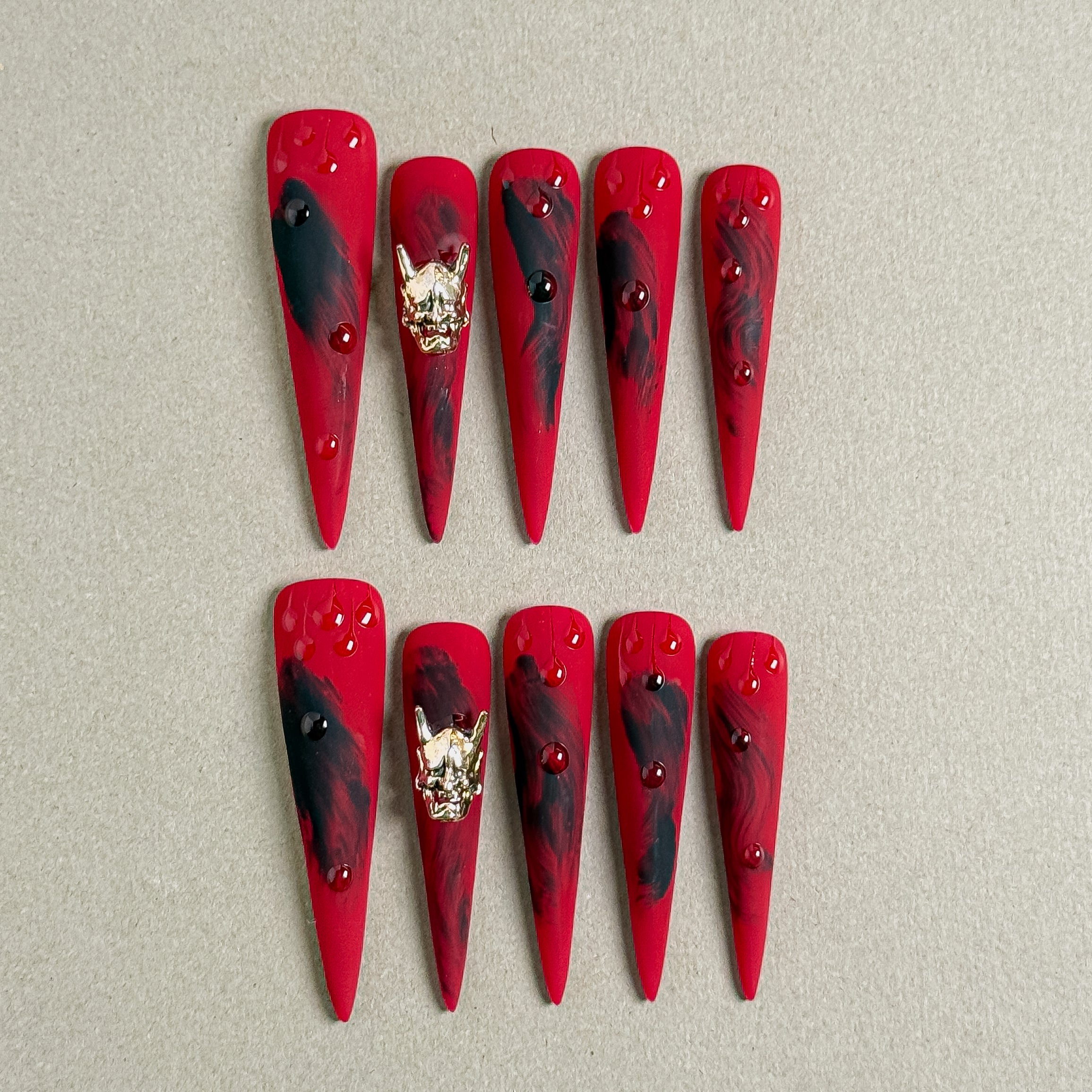 A set of "Fire" stiletto - shaped press - on nails in a vivid red color, adorned with black brush - stroke designs, gold accents, and red rhinestones, ideal for making a striking statement at parties with edgy nail art.
