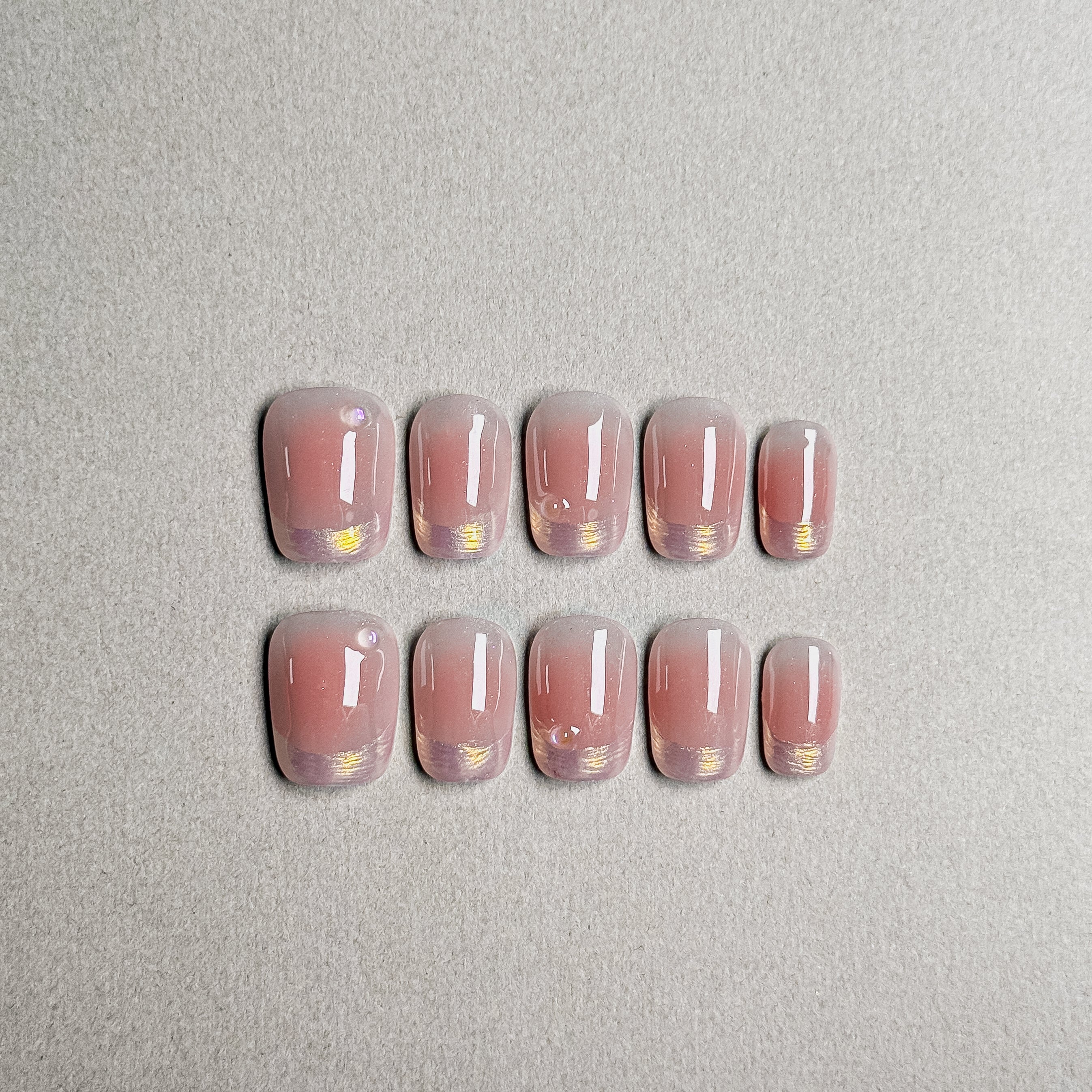 A set of "Sunset" press - on nails featuring a soft gradient of warm pink tones, with delicate gold accents and small embellishments at the base, ideal for achieving a sophisticated everyday nail look.