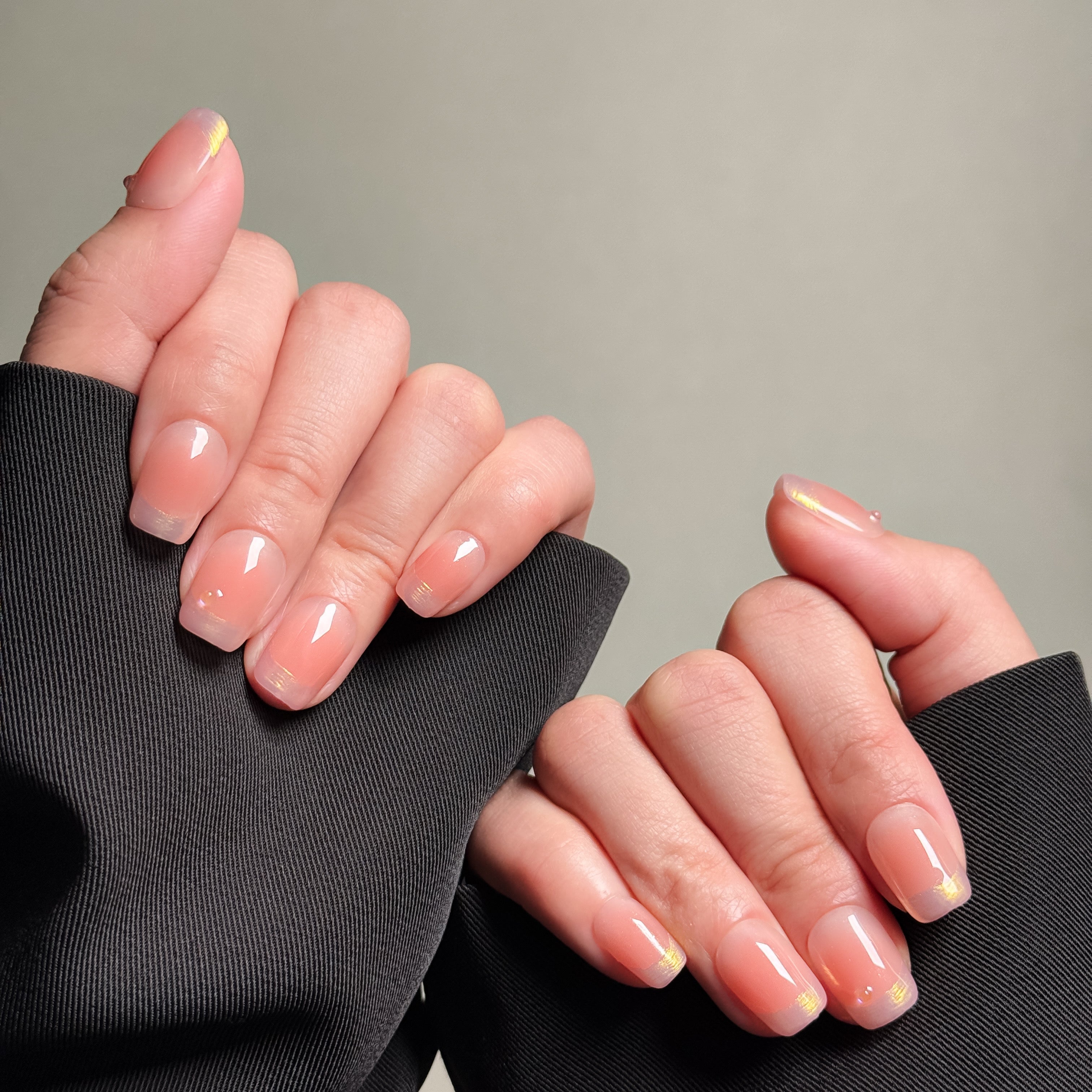 Close-up of hands wearing MIBLAB's nude shimmer press-on nails, showcasing a pearlescent finish with a subtle nude hue. Presented against a gray background and dark fabric, highlighting the delicate shimmer and natural look. These fake nails offer a quick, elegant solution for achieving a natural looking nails effect, perfect for everyday nails or minimalist nails.