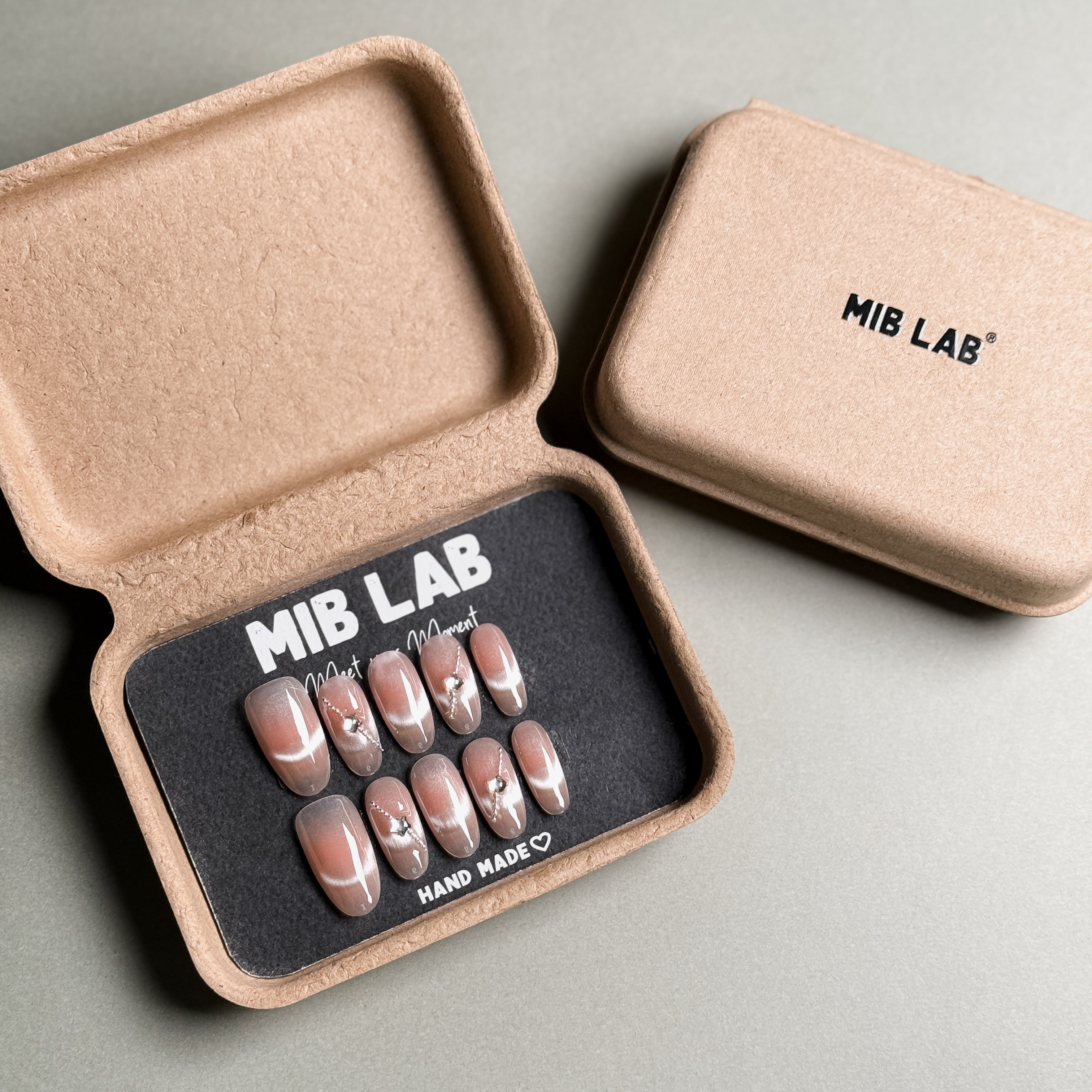 A set of Handmade press on nails with a nude base and delicate silver accents, presented in eco-friendly packaging. These Natural looking nails offer a clean and elegant look, perfect for Everyday nails and professional settings. The box, labeled "MIB LAB" and "Meet Your Moment", highlights the brand's dedication to creating personalized Nail art designs. Ideal for those seeking Reusable fake nails that combine style and sustainability.