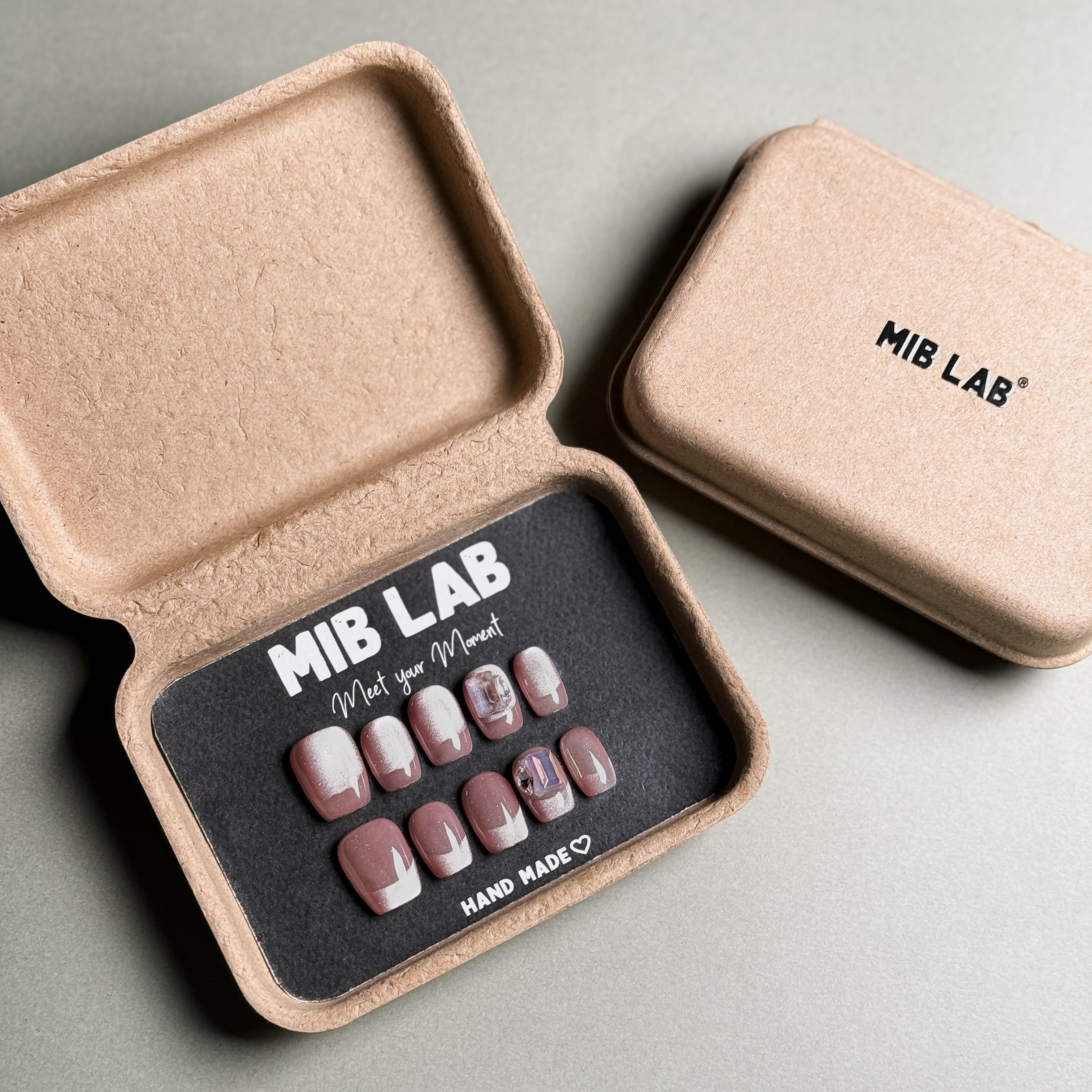 MIBLAB brand pink glitter French tip press-on nails set featuring a dazzling shimmer effect with a subtle pink hue and a classic French tip nails design, packaged in an eco-friendly recycled paper box with the brand name "MIB LAB" and the slogan "Meet your Moment" displayed, along with the "HAND MADE ♡" inscription, presented against a neutral gray background, showcasing short nails design. 