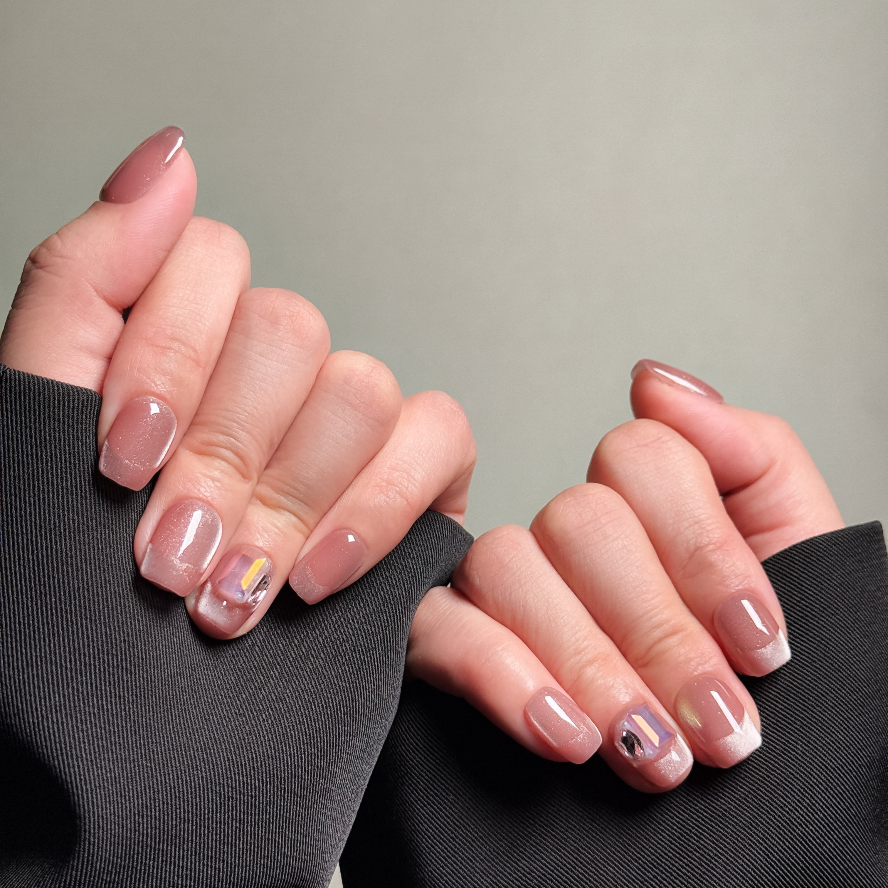 Close-up of hands wearing MIBLAB's pink glitter French tip press-on nails, featuring a shimmering pink hue and classic French tip nails with abstract accents. Presented against a gray background and dark fabric, highlighting the elegant nail art design. These fake nails offer a quick, luxurious press-on nails experience, perfect for everyday nails or party nails.