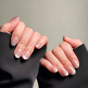 Close-up shot of hands wearing MIBLAB's silver cat eye French tip press-on nails, featuring a mesmerizing shimmering effect with a subtle silver hue and a classic French tip nails design, presented against a neutral gray background and a dark fabric texture, highlighting the glamorous look and polished finish on both short and medium nail lengths, showcasing short nails design. These fake nails offer a quick and easy solution for achieving a sophisticated nail art design.
