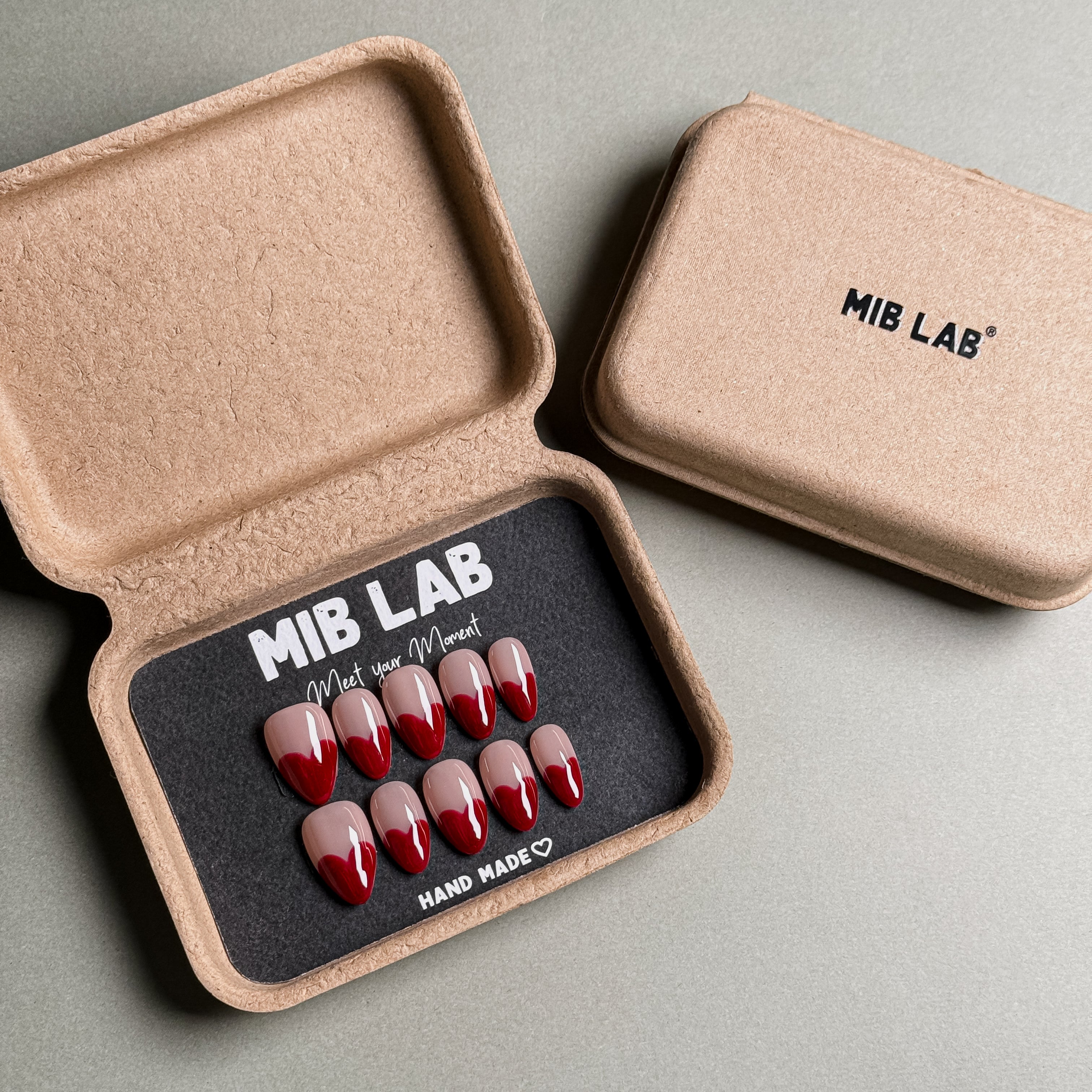 MIBLAB brand red French tip press-on nails set featuring a classic French tip nails design with a bold red tip and a neutral base, packaged in an eco-friendly recycled paper box with the brand name "MIB LAB" and the slogan "Meet your Moment" displayed, along with the "HAND MADE ♡" inscription, presented against a neutral gray background, showcasing short nails design. These fake nails offer a quick and easy solution for achieving a timeless nail art design, suitable for everyday nails or wedding nails.