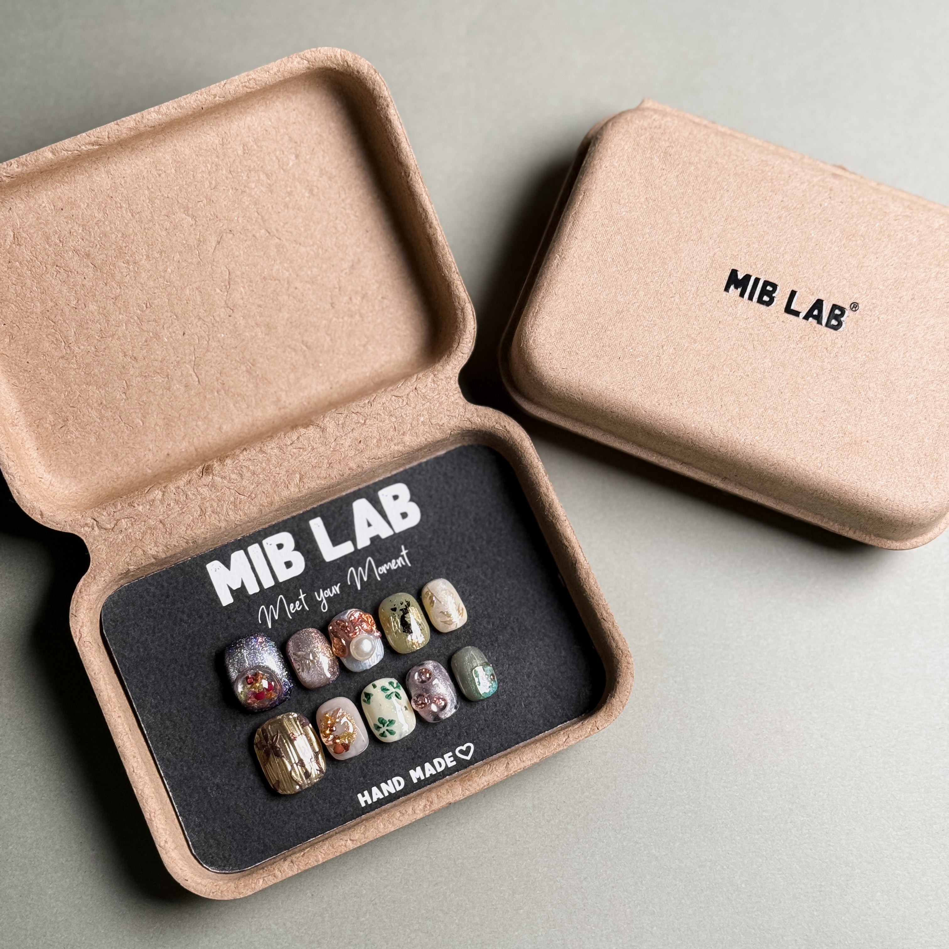 A set of handcrafted Handmade press on nails displayed in a sustainable packaging box, featuring a variety of unique designs. This Nail art kit showcases Custom press on nails with intricate details and embellishments. Ideal for Nail art designs and those seeking Luxury press on nails for special occasions. The box is labeled "MIB LAB" and "Meet Your Moment", emphasizing the brand's commitment to quality and individuality.