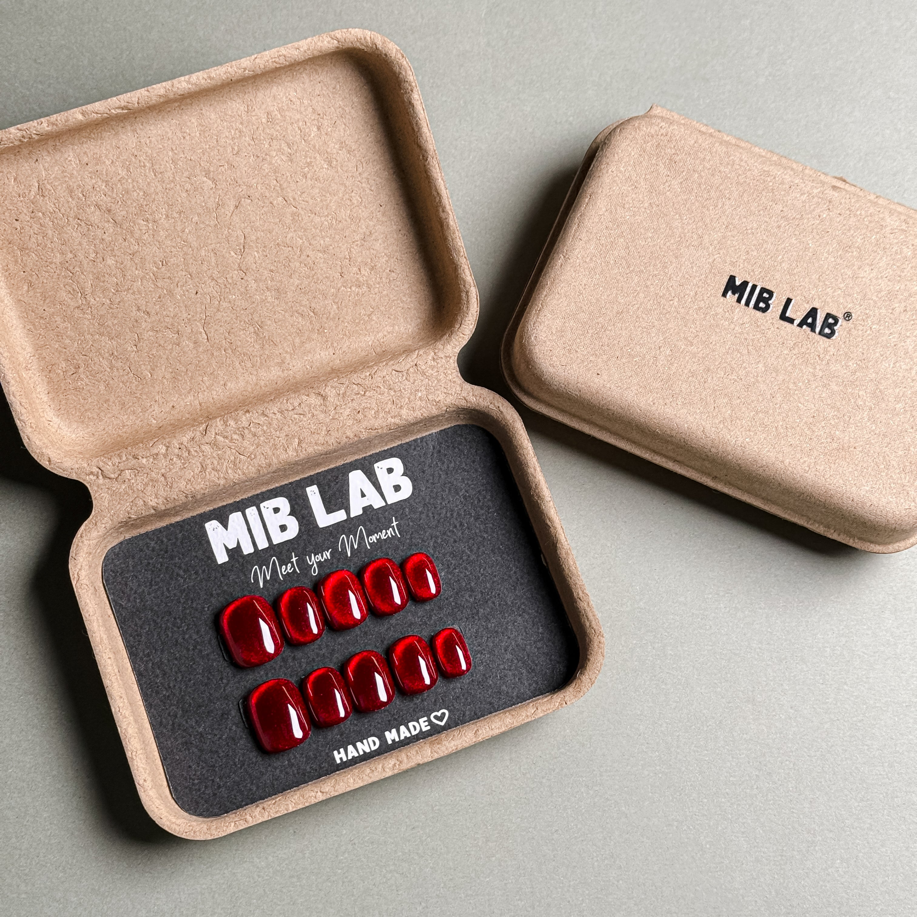 MIBLAB brand red press-on nails set featuring a high-gloss finish, packaged in an eco-friendly recycled paper box with the brand name "MIB LAB" and the slogan "Meet your Moment" displayed, along with the "HAND MADE ♡" inscription, presented against a neutral gray background, showcasing short nails design. These fake nails offer a quick and easy solution for achieving a bold and glamorous look, suitable for party nails or holiday nails.