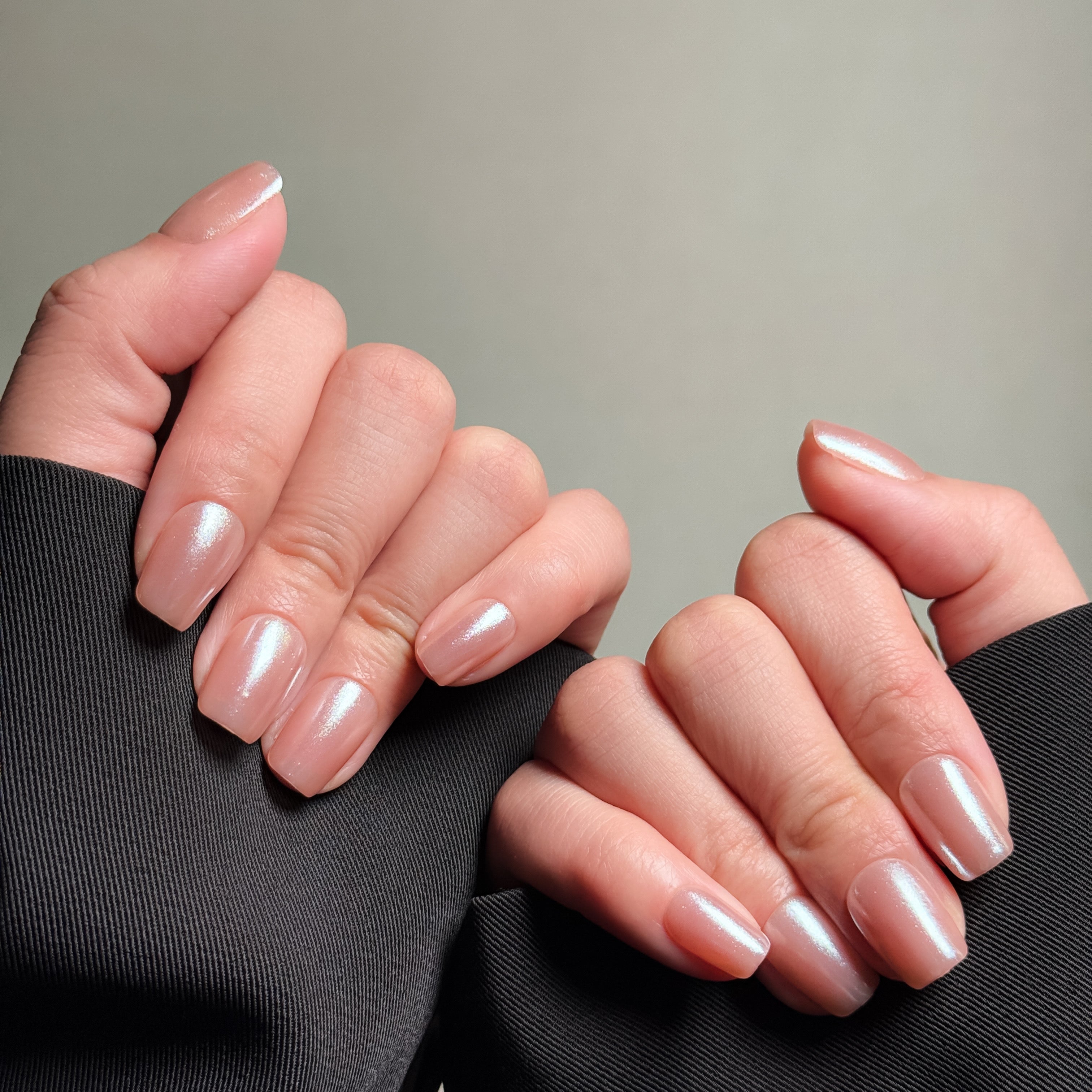 Close-up shot of hands wearing MIBLAB's nude shimmer press-on nails, featuring a pearlescent finish with a subtle nude hue, presented against a neutral gray background and a dark fabric texture, highlighting the delicate shimmer and natural look on both short and medium nail lengths, showcasing short nails design. These fake nails offer a quick and easy solution for achieving a natural looking nails effect with a touch of elegance, suitable for everyday nails or minimalist nails.