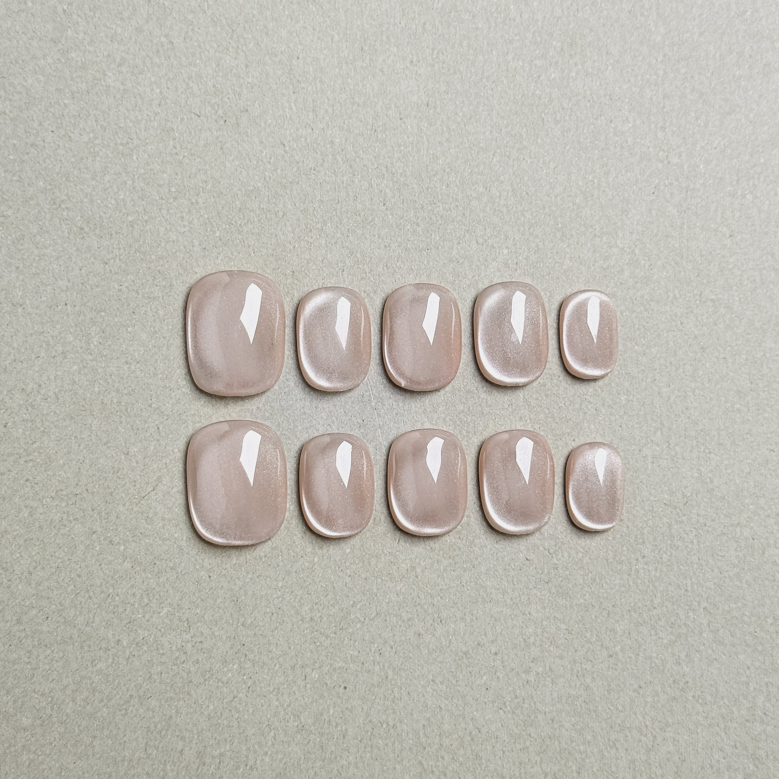 Two rows of MIBLAB's nude cat eye press-on nails, featuring a mesmerizing shimmering effect with a subtle nude hue, arranged against a neutral gray background to highlight the unique texture and depth of the polish, with a gradient of sizes from large to small for a perfect fit, showcasing short nails design. These fake nails offer a quick and easy solution for achieving a natural looking nails effect with a touch of glamour, suitable for everyday nails or minimalist nails.