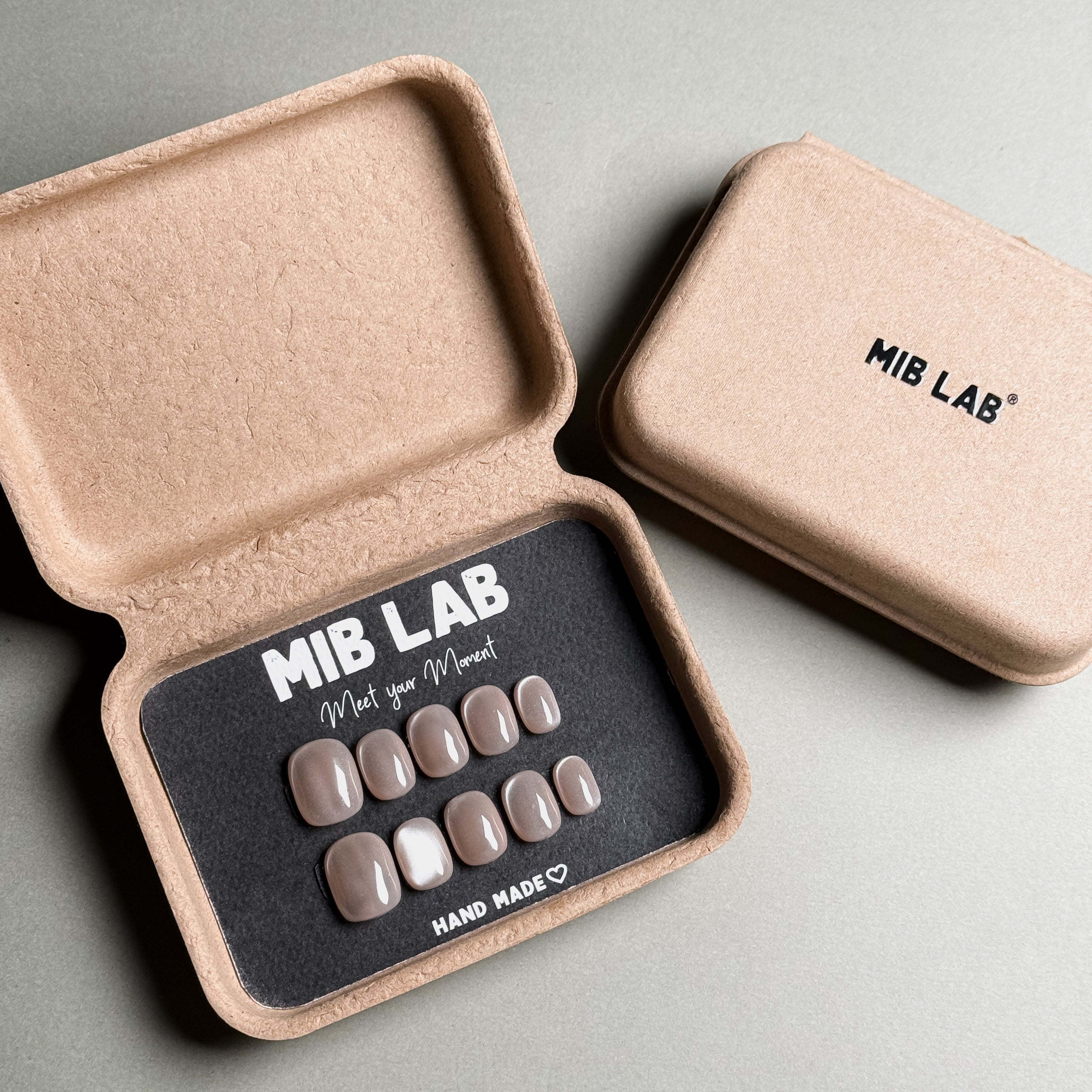 MIBLAB brand nude cat eye press-on nails set featuring a mesmerizing shimmering effect with a subtle nude hue, packaged in an eco-friendly recycled paper box with the brand name "MIB LAB" and the slogan "Meet your Moment" displayed, along with the "HAND MADE ♡" inscription, presented against a neutral gray background, showcasing short nails design. These fake nails offer a quick and easy solution for achieving a natural looking nails effect with a touch of glamour, suitable for everyday nails or minimalist 