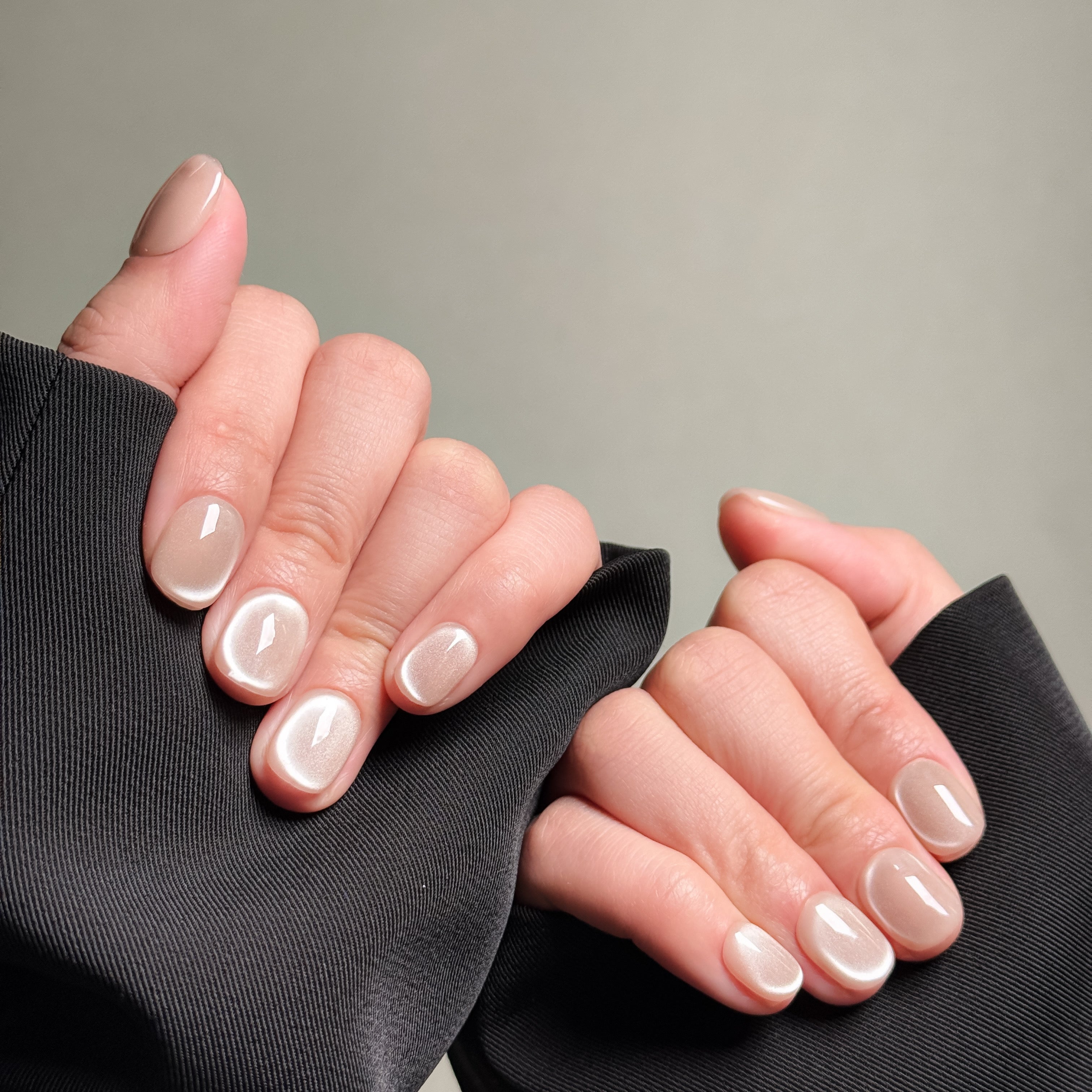 Close-up shot of hands wearing MIBLAB's nude cat eye press-on nails, featuring a mesmerizing shimmering effect with a subtle nude hue, presented against a neutral gray background and a dark fabric texture, highlighting the unique texture and depth of the polish on both short and medium nail lengths, showcasing short nails design. These fake nails offer a quick and easy solution for achieving a natural looking nails effect with a touch of glamour, suitable for everyday nails or minimalist nails.