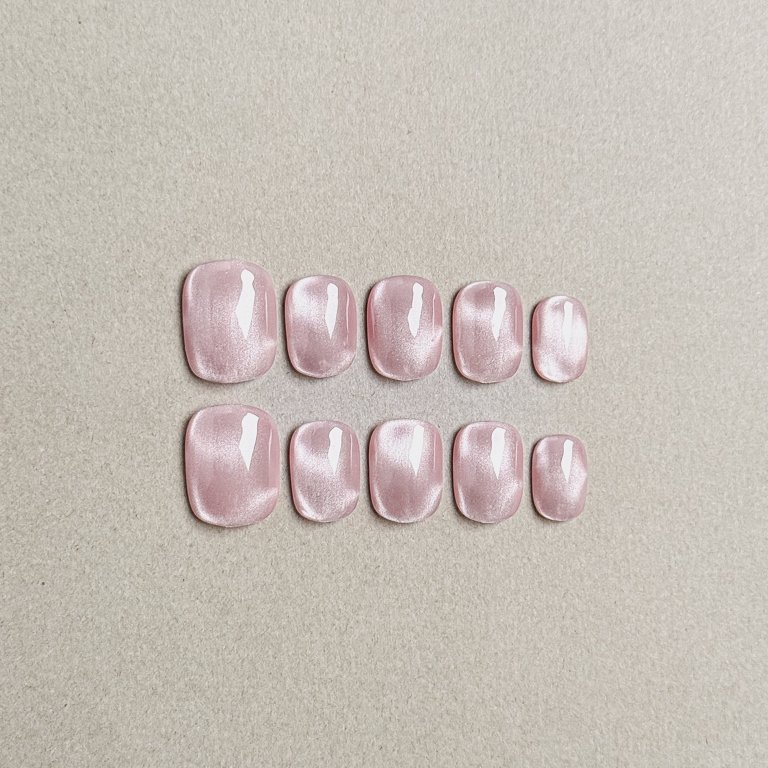 Two rows of MIBLAB's pink cat eye press-on nails, featuring a mesmerizing shimmering effect with a subtle pink hue, arranged against a neutral gray background to highlight the unique texture and depth of the polish, with a gradient of sizes from large to small for a perfect fit, showcasing short nails design. These fake nails offer a quick and easy solution for achieving a natural looking nails effect with a touch of glamour, suitable for everyday nails or party nails.