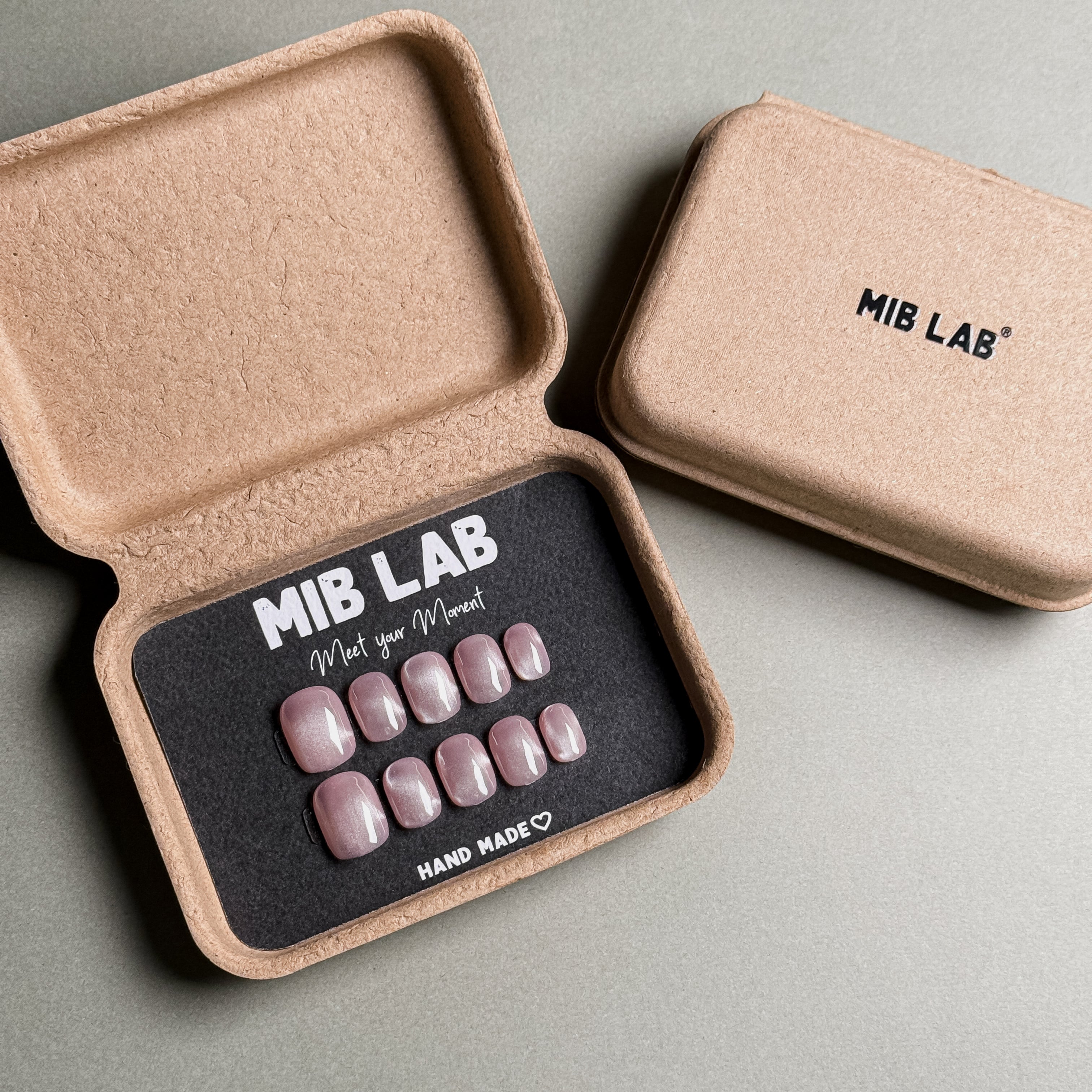 MIBLAB brand pink cat eye press-on nails set featuring a mesmerizing shimmering effect with a subtle pink hue, packaged in an eco-friendly recycled paper box with the brand name "MIB LAB" and the slogan "Meet your Moment" displayed, along with the "HAND MADE ♡" inscription, presented against a neutral gray background, showcasing short nails design. These fake nails offer a quick and easy solution for achieving a natural looking nails effect with a touch of glamour, suitable for everyday nails or party nails