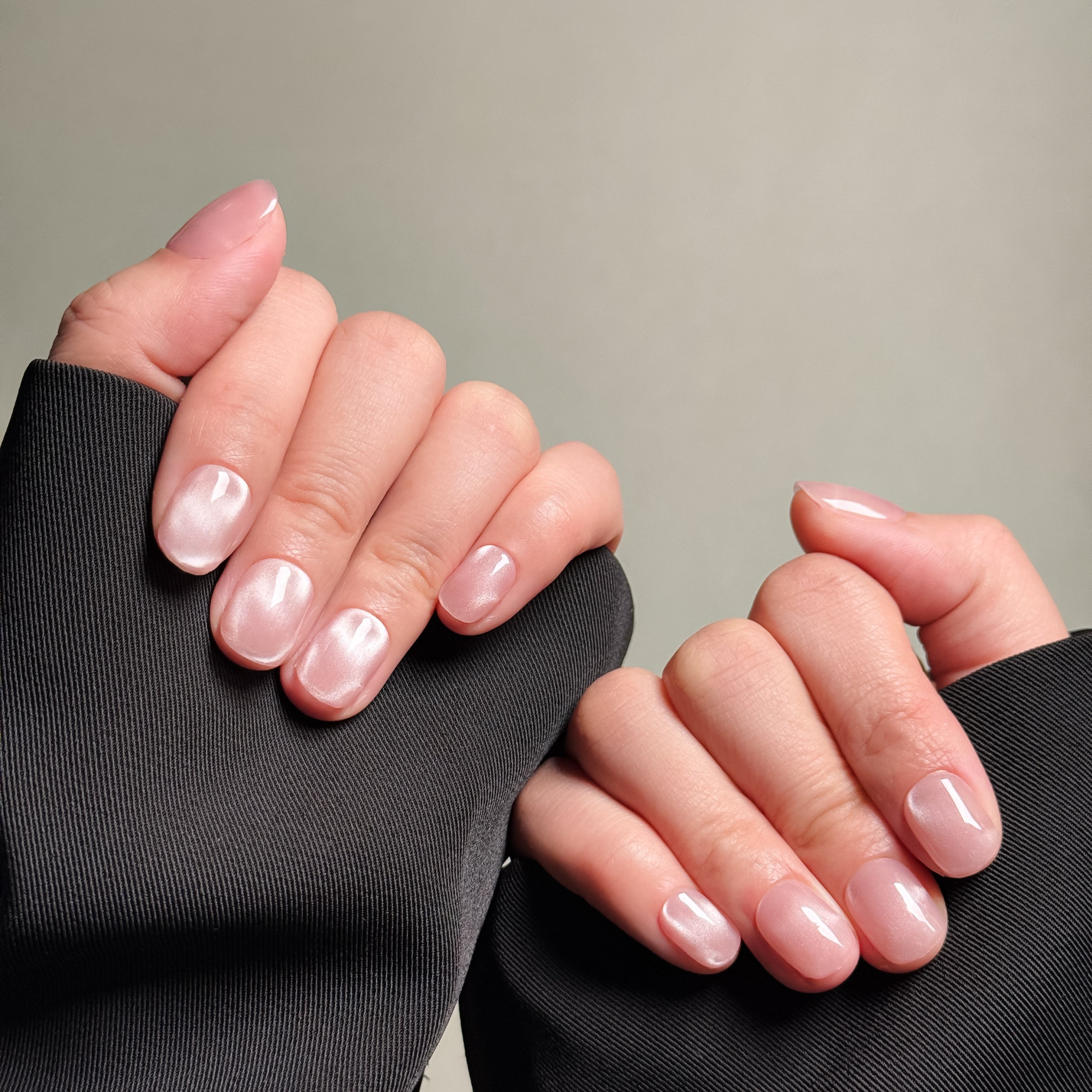 Close-up shot of hands wearing MIBLAB's pink cat eye press-on nails, featuring a mesmerizing shimmering effect with a subtle pink hue, presented against a neutral gray background and a dark fabric texture, highlighting the unique texture and depth of the polish on both short and medium nail lengths, showcasing short nails design. These fake nails offer a quick and easy solution for achieving a natural looking nails effect with a touch of glamour, suitable for everyday nails or party nails.