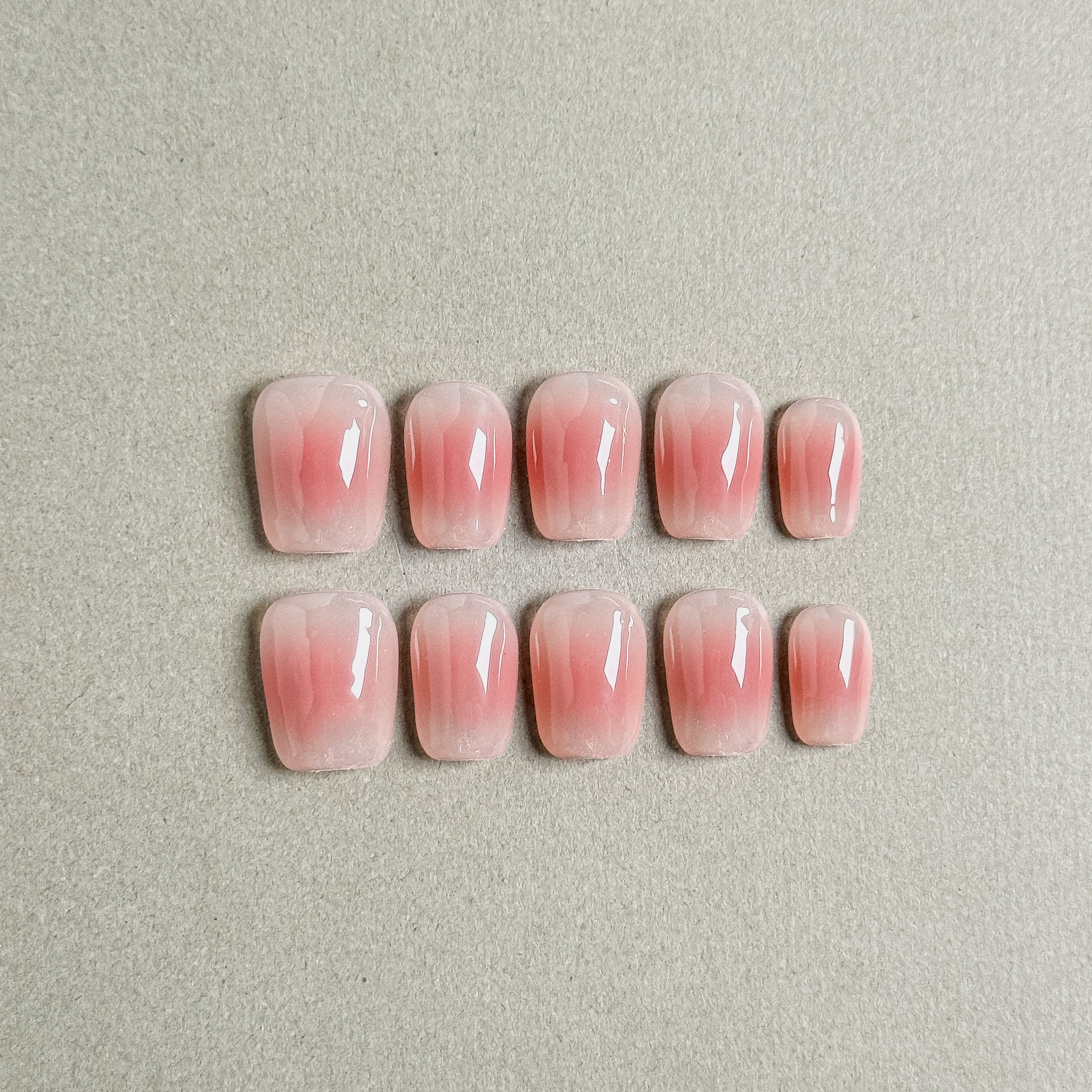 Two rows of MIBLAB's pink press-on nails featuring a delicate French tip nails design with a soft pink gradient effect, arranged against a neutral gray background to highlight the subtle color transition and elegant style, with a gradient of sizes from large to small for a perfect fit, showcasing short nails design. These fake nails offer a quick and easy solution for achieving a natural looking nails effect, suitable for everyday nails or as wedding nails.