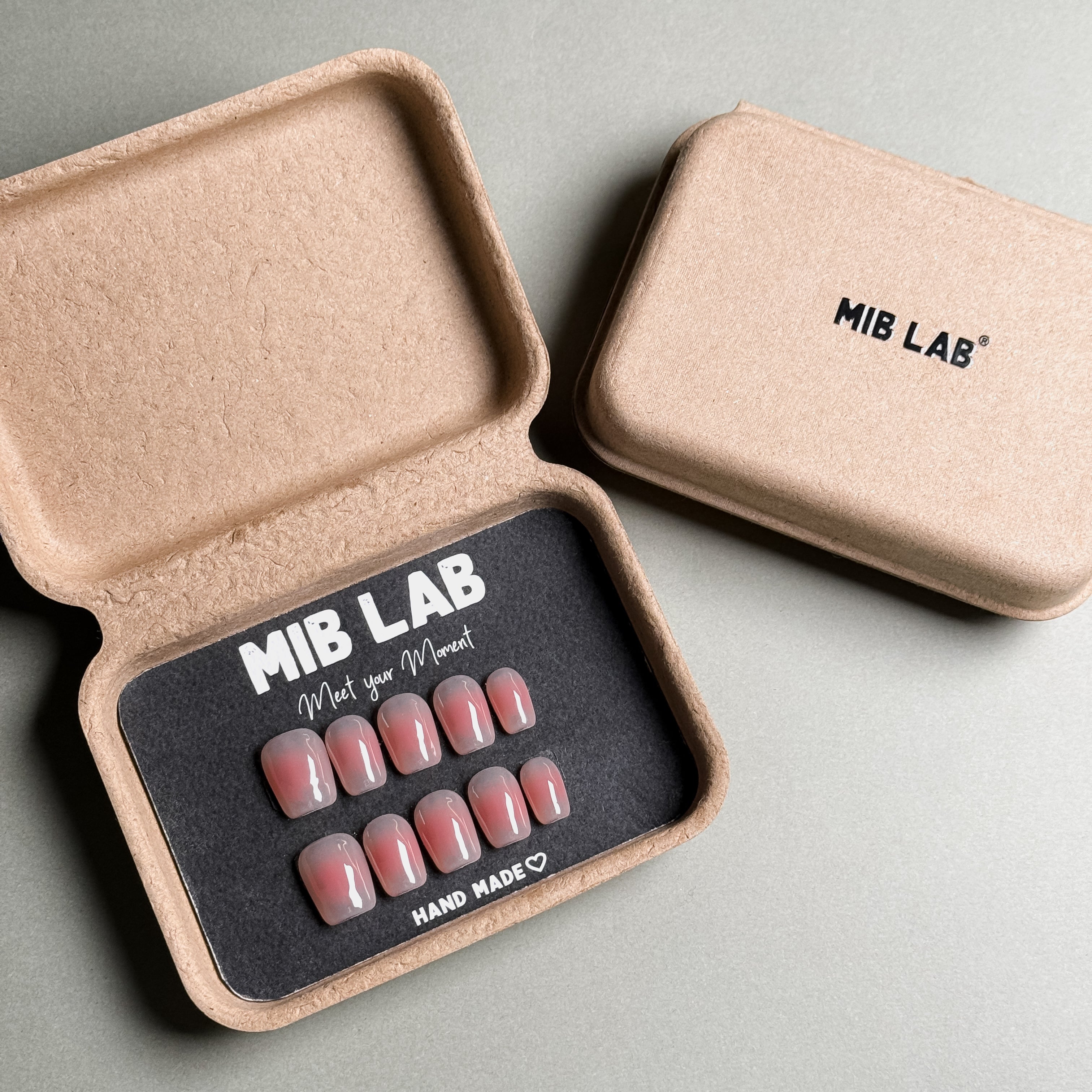 MIBLAB brand pink press-on nails set featuring a delicate French tip nails design with a soft pink gradient effect, packaged in an eco-friendly recycled paper box with the brand name "MIB LAB" and the slogan "Meet your Moment" displayed, along with the "HAND MADE ♡" inscription, presented against a neutral gray background, showcasing short nails design. These fake nails offer a quick and easy solution for achieving a natural looking nails effect, suitable for everyday nails or as wedding nails.