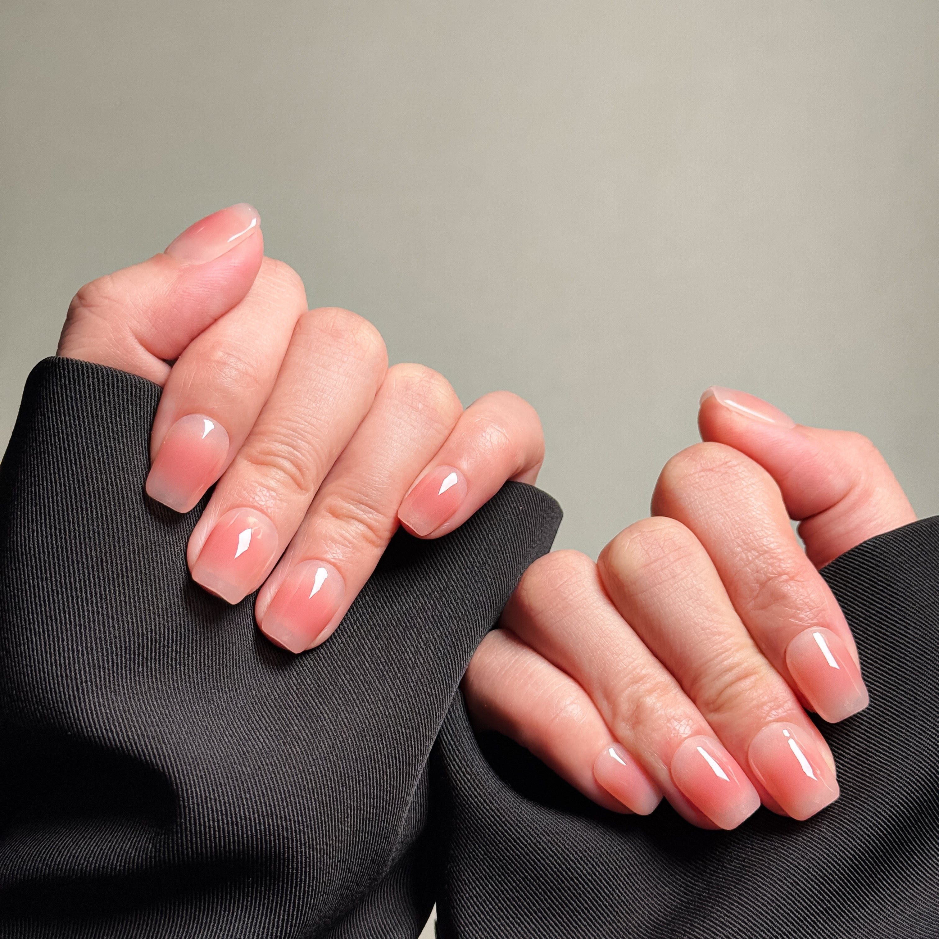 Close-up shot of hands wearing MIBLAB's pink press-on nails featuring a delicate French tip nails design with a soft pink gradient effect, presented against a neutral gray background and a dark fabric texture, highlighting the elegant and polished look of the nail art design. These fake nails offer a quick and easy solution for achieving short nails design with a natural looking nails effect, suitable for everyday nails or as wedding nails.