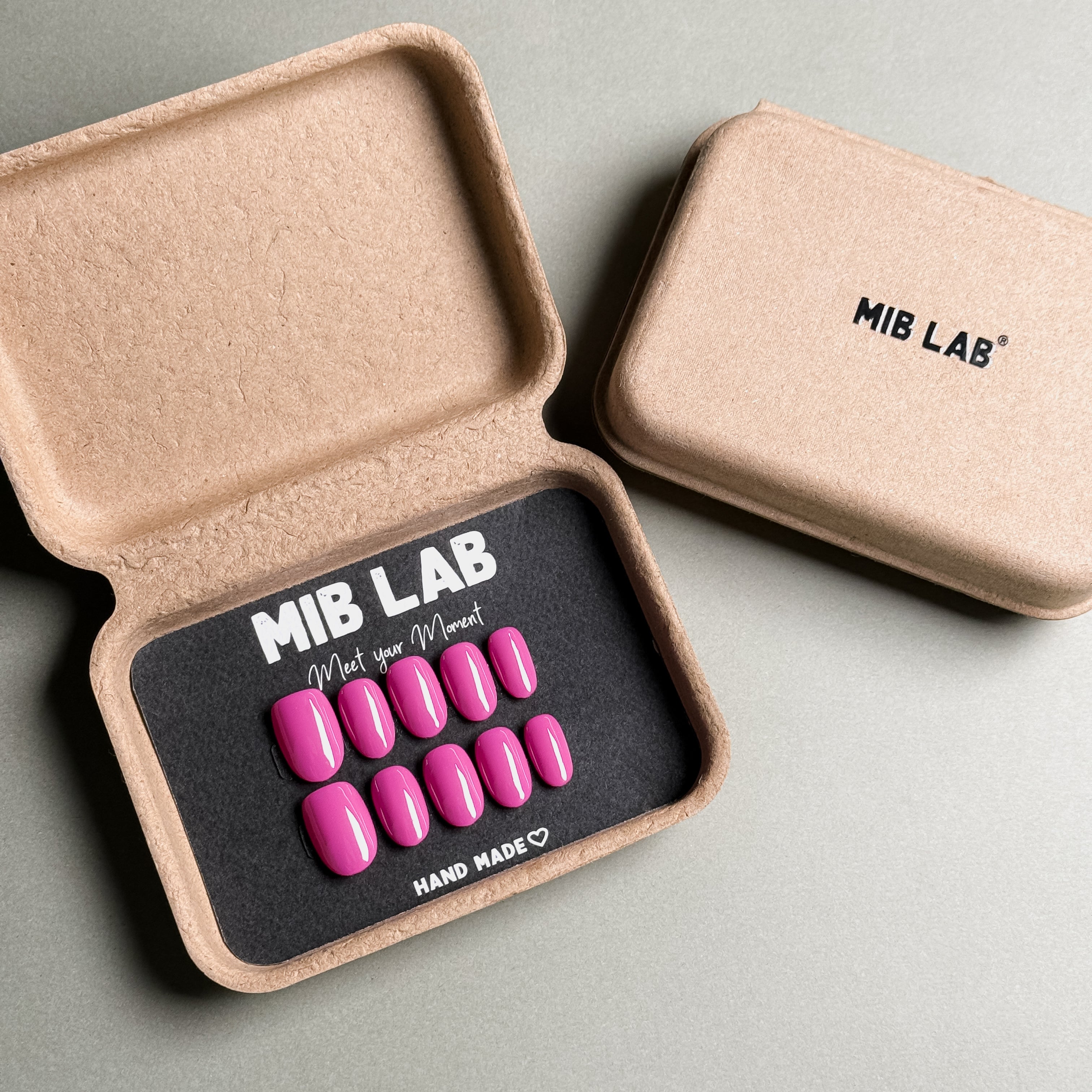MIBLAB brand glue-on nails set featuring vibrant pink fake nails with a high-gloss finish, packaged in an eco-friendly recycled paper box with the brand name "MIB LAB" and the slogan "Meet your Moment" displayed, along with the "HAND MADE ♡" inscription, presented against a neutral gray background, showcasing a short nails design option. These reusable fake nails are perfect for creating everyday nails or party nails looks, offering a quick and easy nail art solution with a natural looking nails effect.