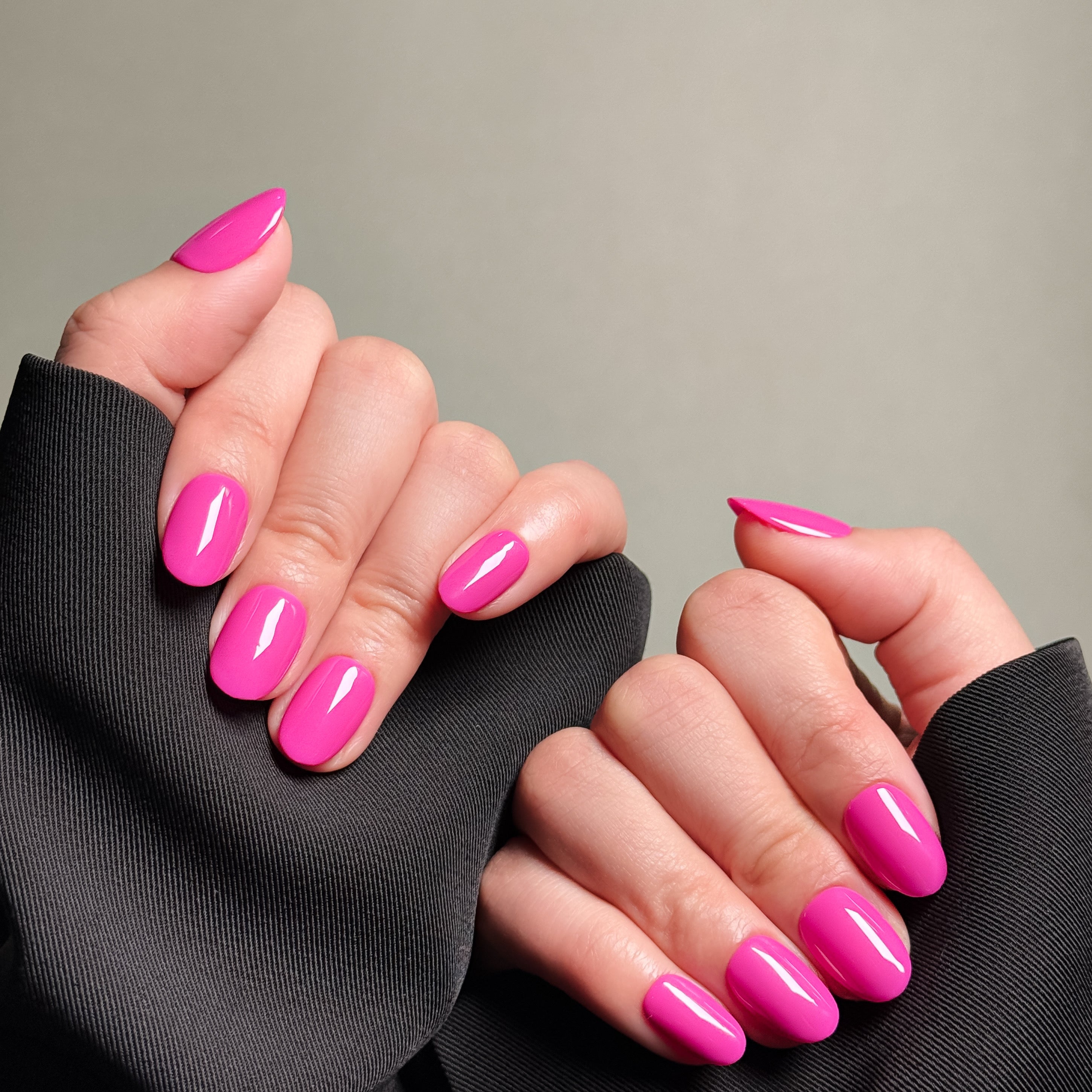 Close-up shot of hands wearing MIBLAB's pink press-on nails in a stylish almond nails shape, featuring a high-gloss finish that enhances the vibrant color, presented against a neutral gray background and a dark fabric texture, highlighting the elegant and polished look of the nail art design. These fake nails offer a quick and easy solution for achieving long nails design with a natural looking nails effect, suitable for everyday nails or party nails.