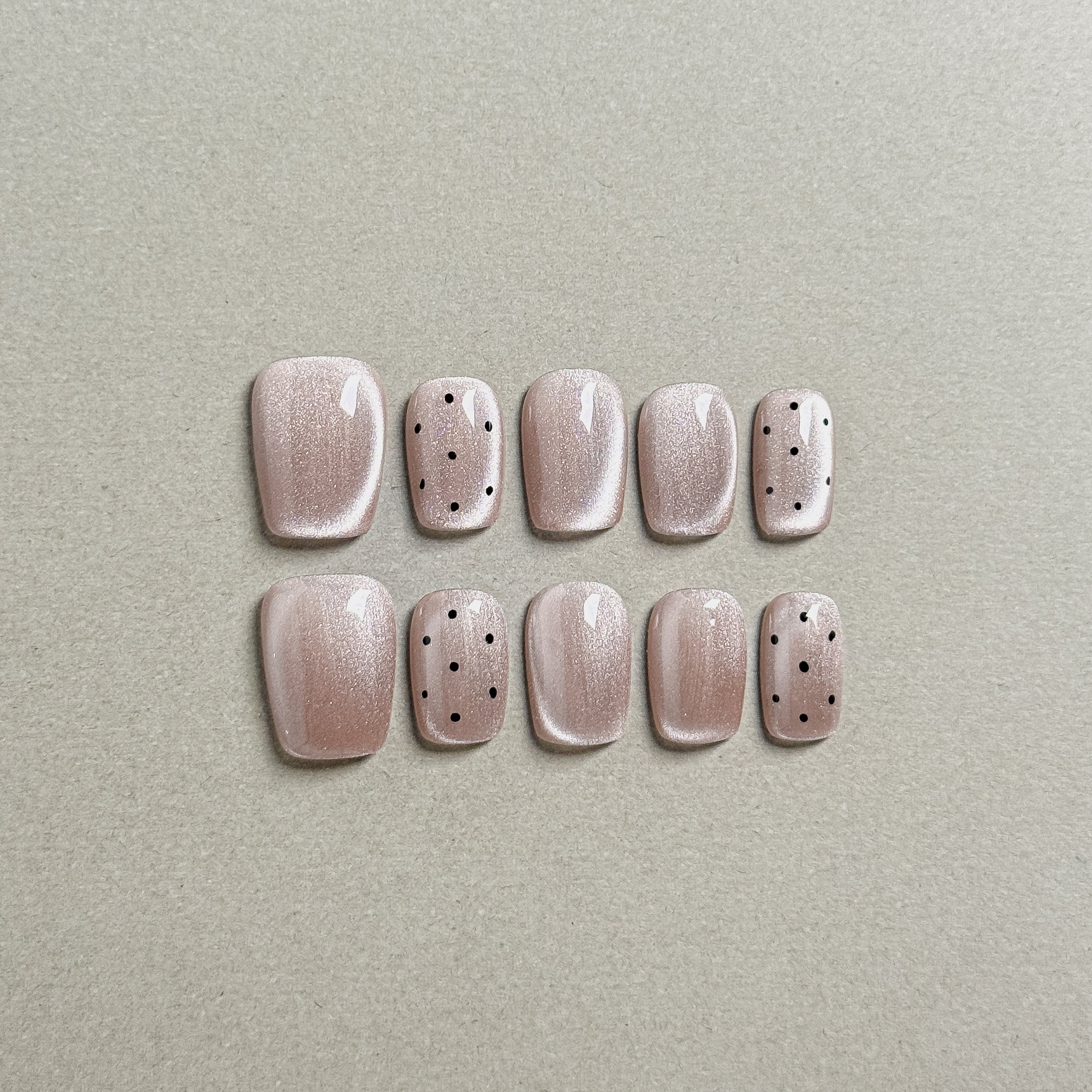 Two rows of MIBLAB's light pink press-on toenails with subtle black polka dot designs, featuring a soft shimmer and a glossy finish, arranged against a neutral gray background to highlight the delicate pattern and elegant color palette, with a gradient of sizes from large to small for a perfect fit.