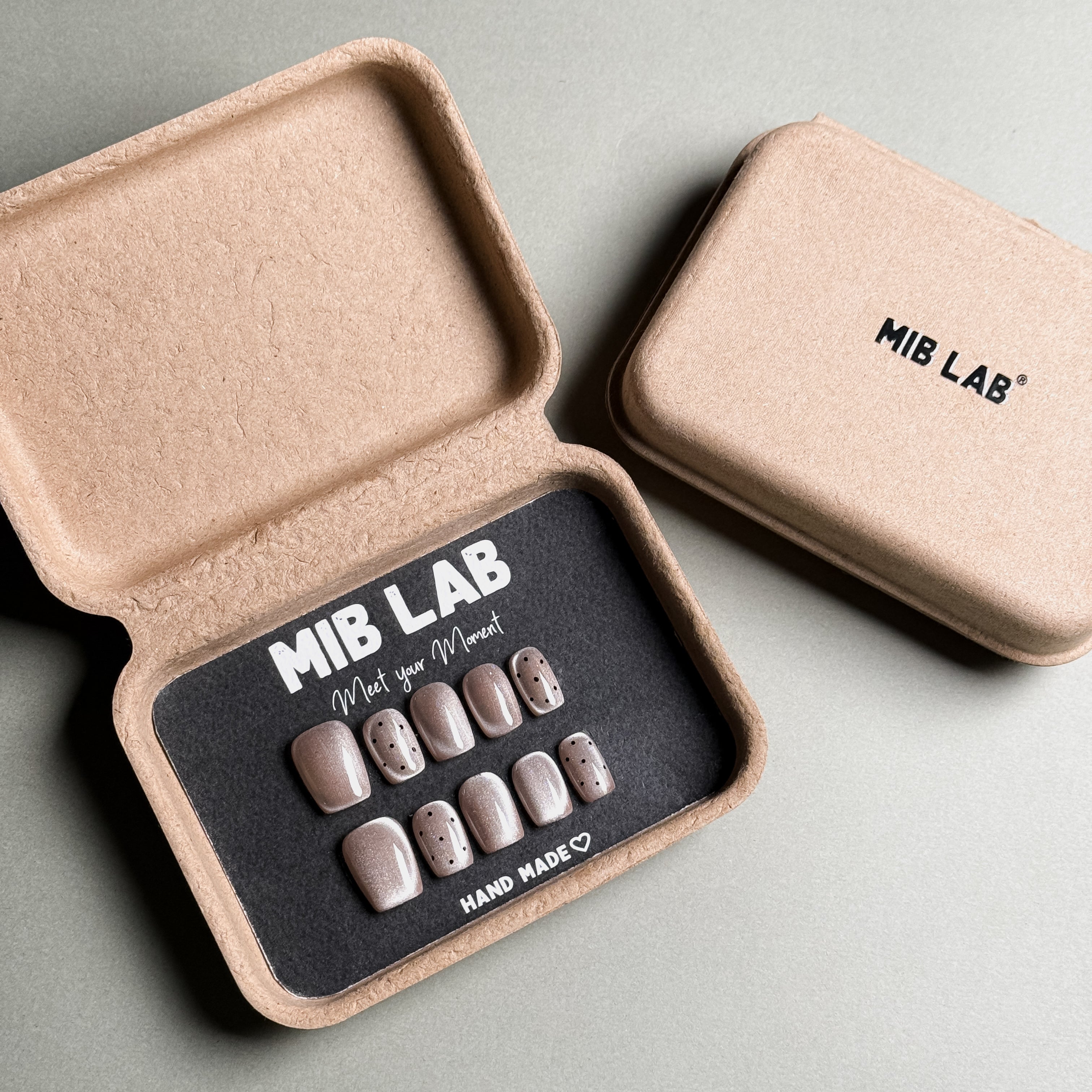 MIBLAB brand press-on toenail set featuring light pink toenails with subtle black polka dot designs and a soft shimmer, packaged in an eco-friendly recycled paper box with the brand name "MIB LAB" and the slogan "Meet your Moment" displayed, along with the "HAND MADE ♡" inscription, presented against a neutral gray background.
