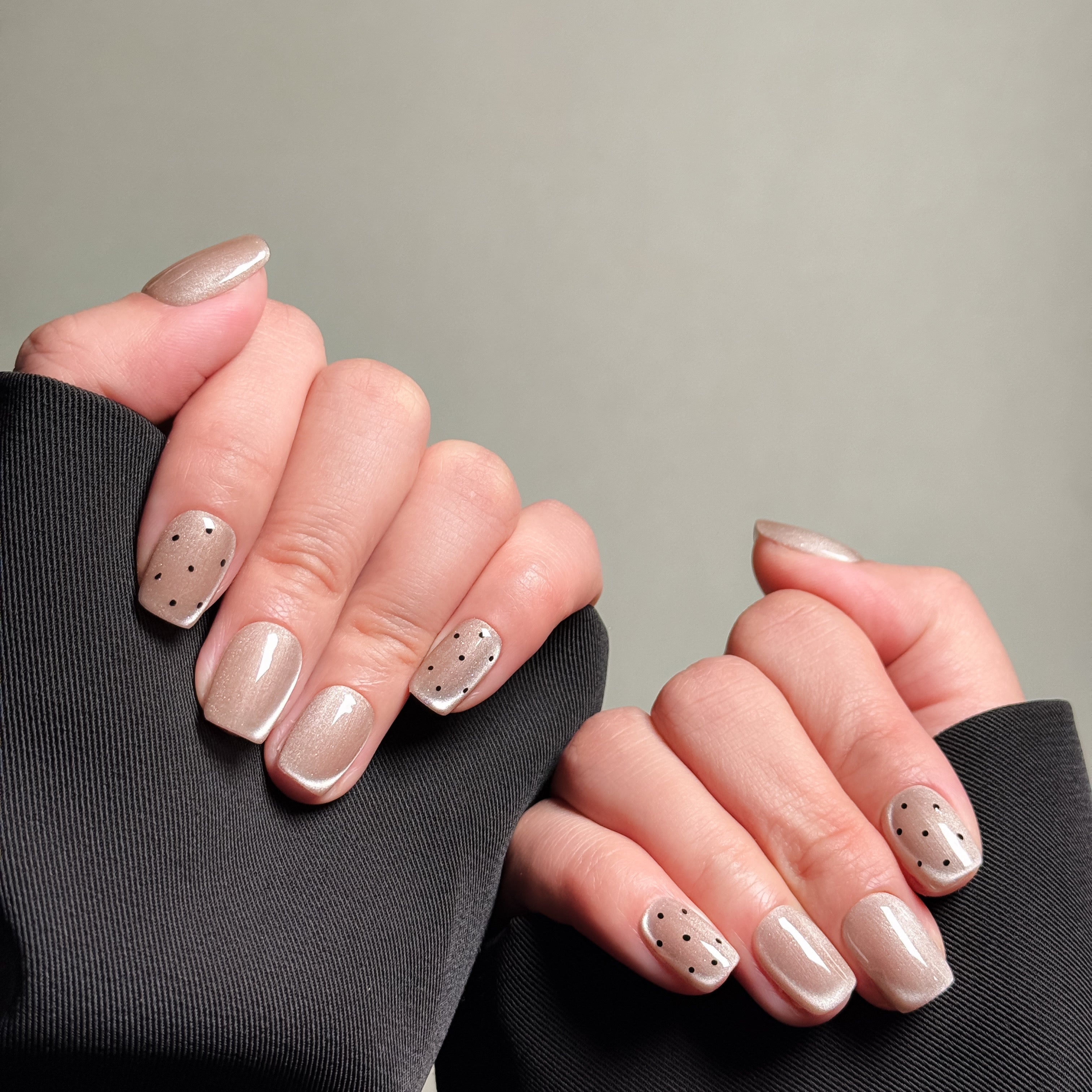Close-up shot of hands wearing MIBLAB's light pink press-on nails with subtle black polka dot designs, featuring a soft shimmer and a glossy finish, presented against a neutral gray background and a black fabric texture, highlighting the delicate pattern and elegant color palette on both short and long nail shapes.