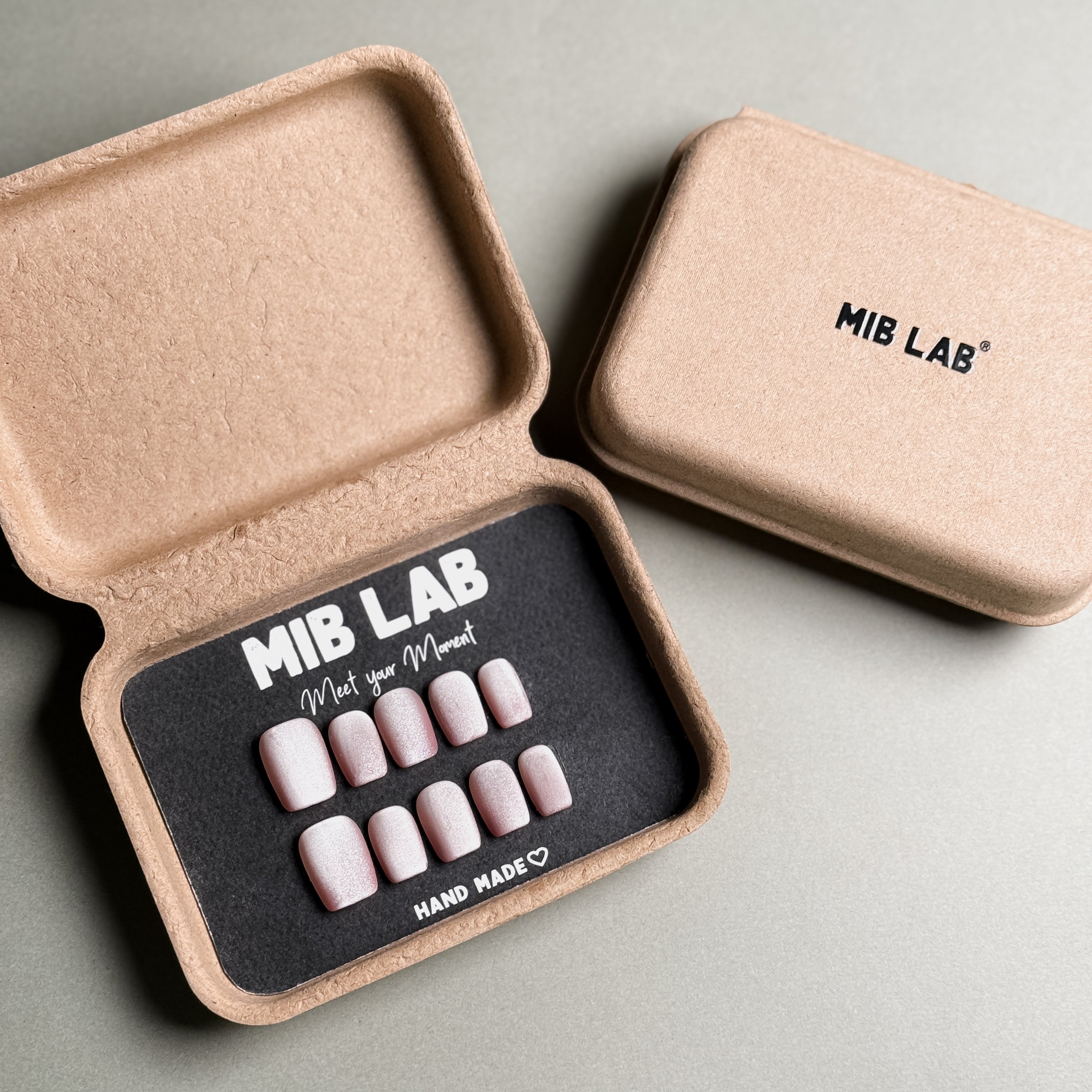 MIBLAB brand press-on toenail set featuring light pink toenails with delicate pink floral designs and a subtle shimmer, packaged in an eco-friendly recycled paper box with the brand name "MIB LAB" and the slogan "Meet your Moment" displayed, along with the "HAND MADE ♡" inscription, presented against a neutral gray background.