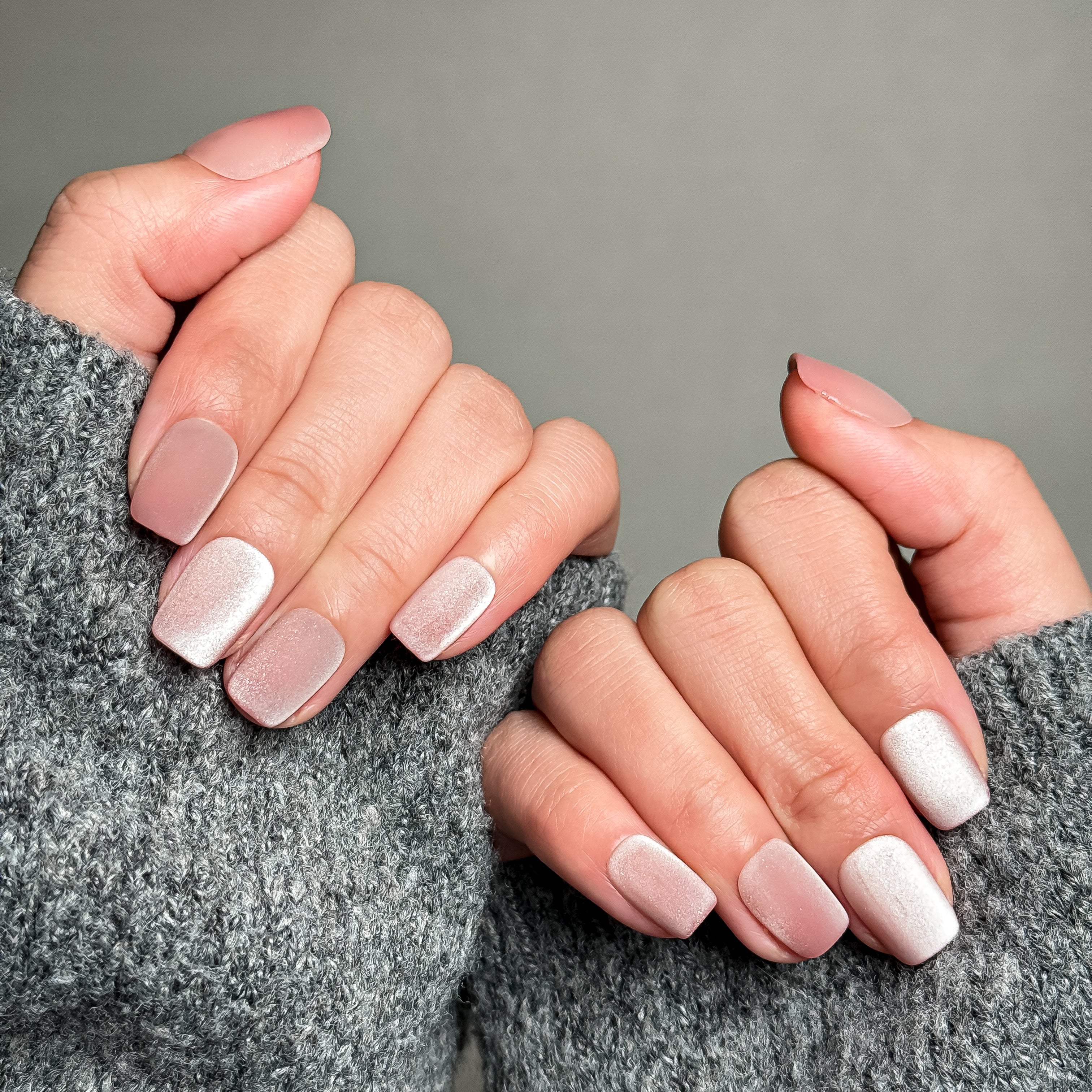 Close-up shot of hands wearing MIBLAB's soft pink gradient press-on nails, showcasing a smooth transition from a matte light pink to a shimmering white, presented against a neutral gray background and a gray knitted sweater, highlighting the subtle color blend and the delicate texture of the manicure.