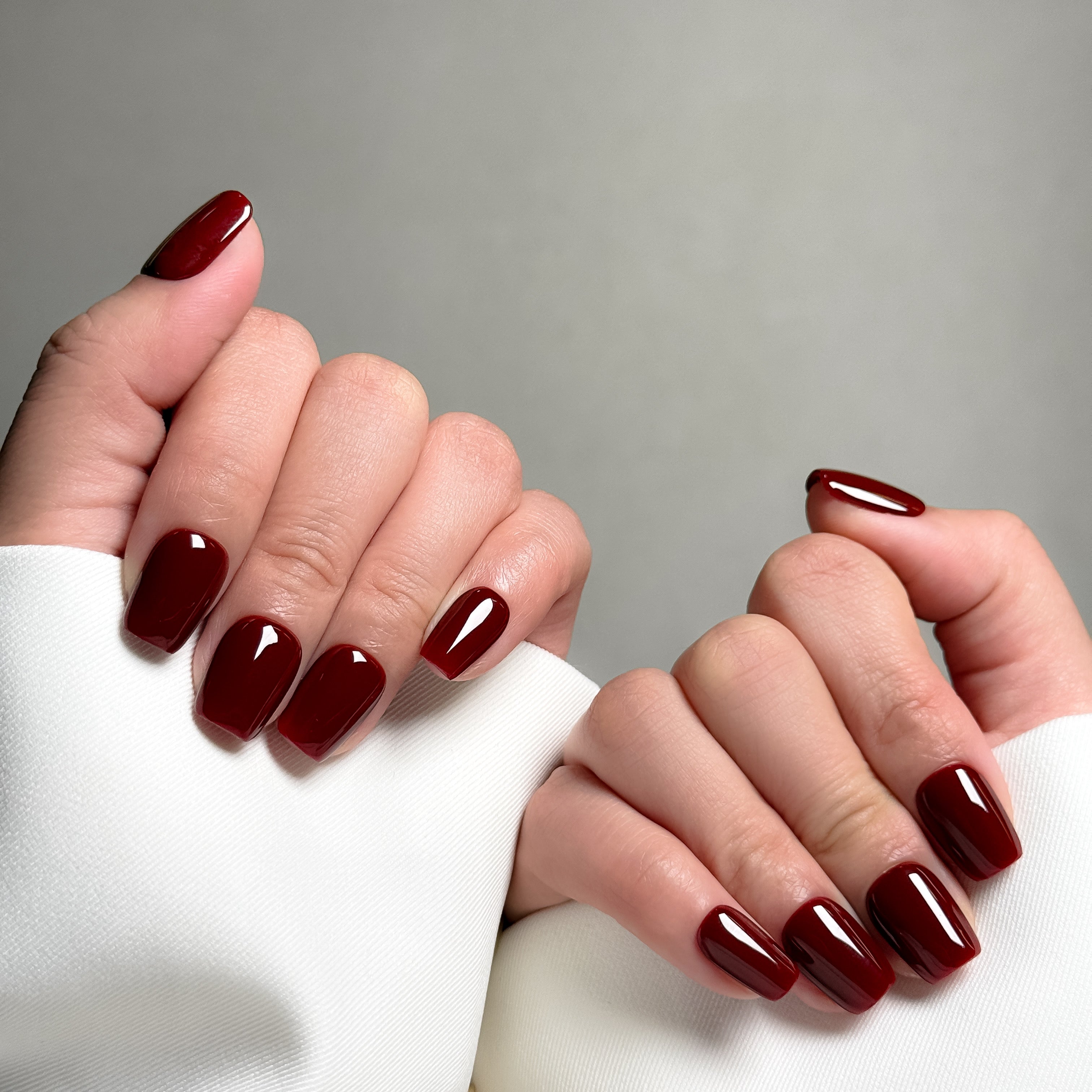 Close-up shot of hands wearing MIBLAB's elegant wine red press-on nails in a classic coffin shape, featuring a high-gloss finish that enhances the rich, deep color, presented against a neutral gray background and a white fabric texture, highlighting the sophisticated and polished look of the manicure.