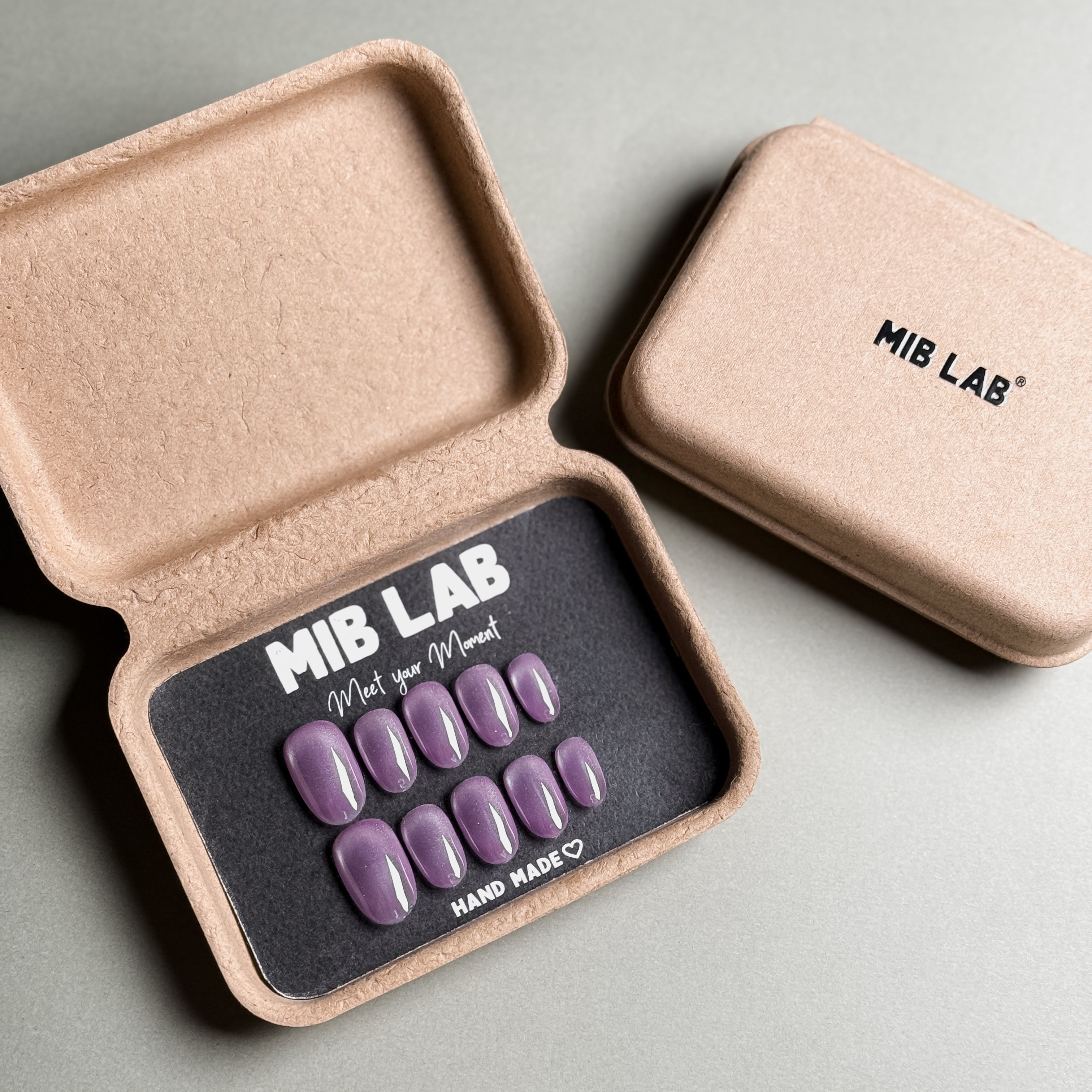 A set of Handmade press on nails with a solid purple color, presented in eco-friendly packaging. These Minimalist nails offer a clean and sophisticated look, perfect for Everyday nails and professional settings. The box, labeled "MIB LAB" and "Meet Your Moment", highlights the brand's dedication to creating timeless Nail art designs. Ideal for those seeking Reusable fake nails that are both stylish and sustainable.