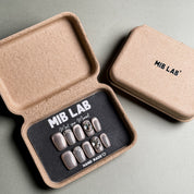 A set of Handmade press on nails with a metallic gray finish and rhinestone accents, presented in eco-friendly packaging. These Luxury press on nails offer a glamorous and eye-catching look, perfect for Party nails or special occasions. The box, labeled "MIB LAB" and "Meet Your Moment", highlights the brand's dedication to creating memorable Nail art designs. Ideal for those seeking Reusable fake nails that combine style and sustainability.