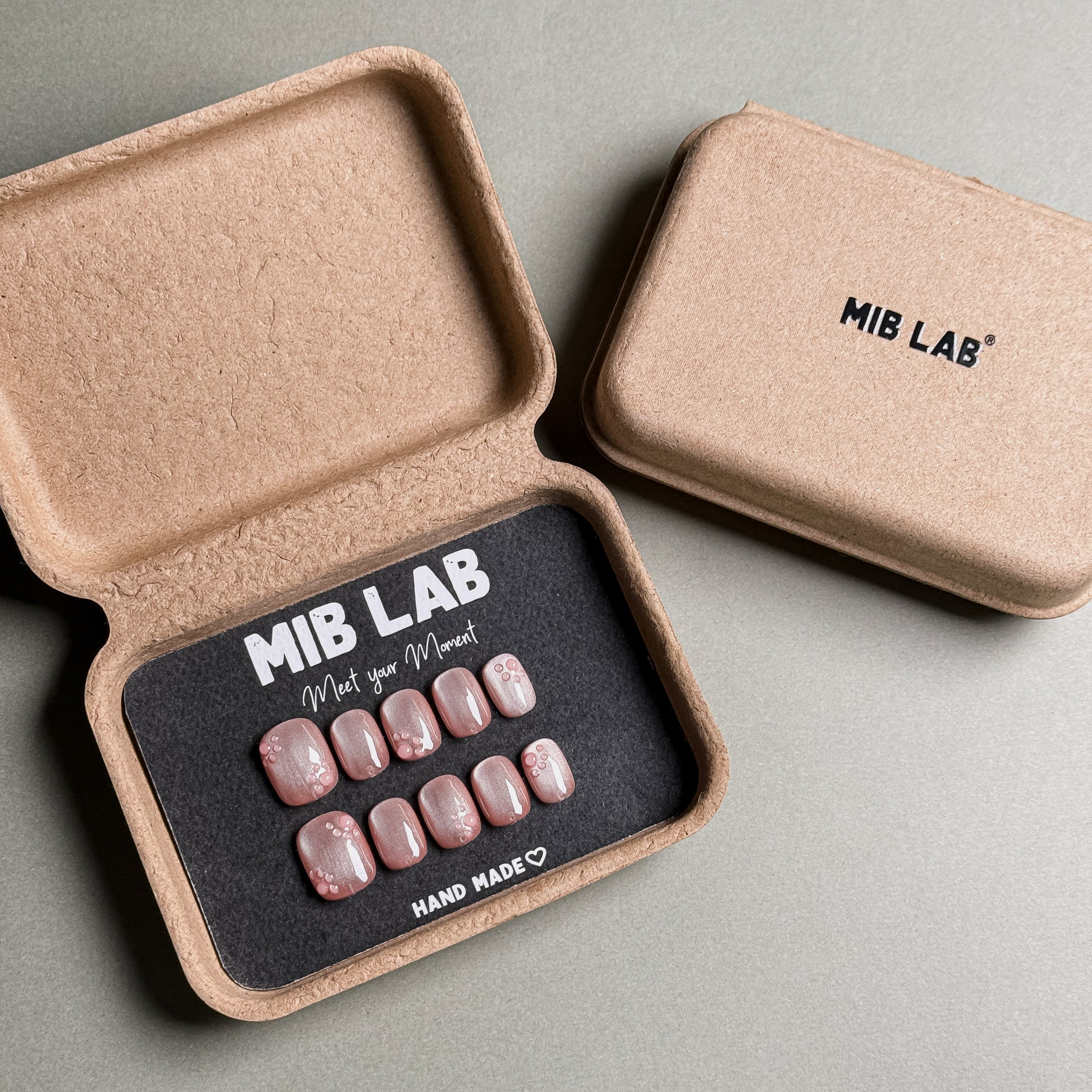 MIBLAB brand press-on toenail set featuring light pink toenails with delicate pink floral designs and a subtle shimmer, packaged in an eco-friendly recycled paper box with the brand name "MIB LAB" and the slogan "Meet your Moment" displayed, along with the "HAND MADE ♡" inscription, presented against a neutral gray background.