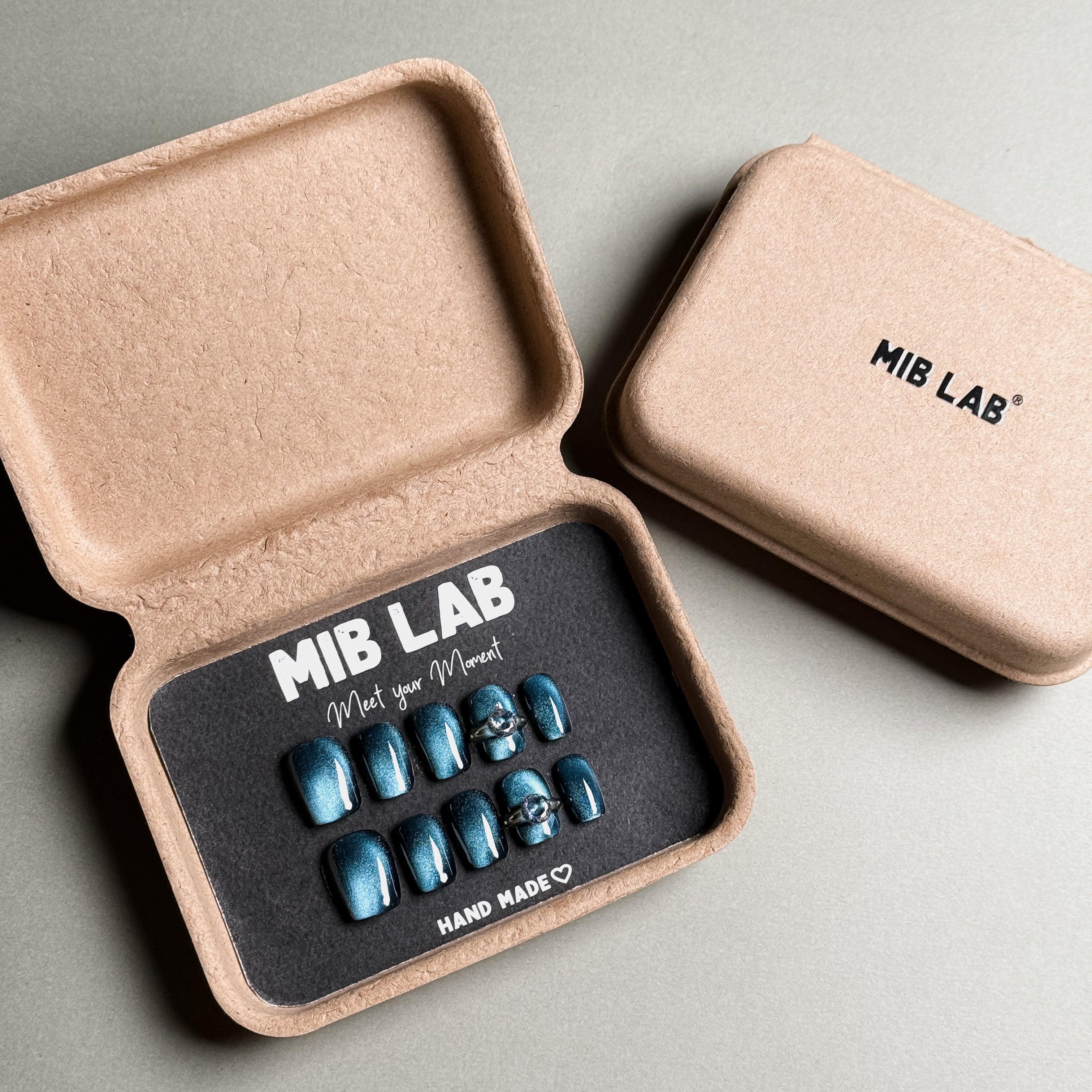 A set of Handmade press on nails with a deep teal finish and silver ring accents, presented in eco-friendly packaging. These Luxury press on nails offer a bold and unique look, perfect for Party nails or special occasions. The box, labeled "MIB LAB" and "Meet Your Moment", highlights the brand's dedication to creating memorable Nail art designs. Ideal for those seeking Reusable fake nails that combine style and sustainability.